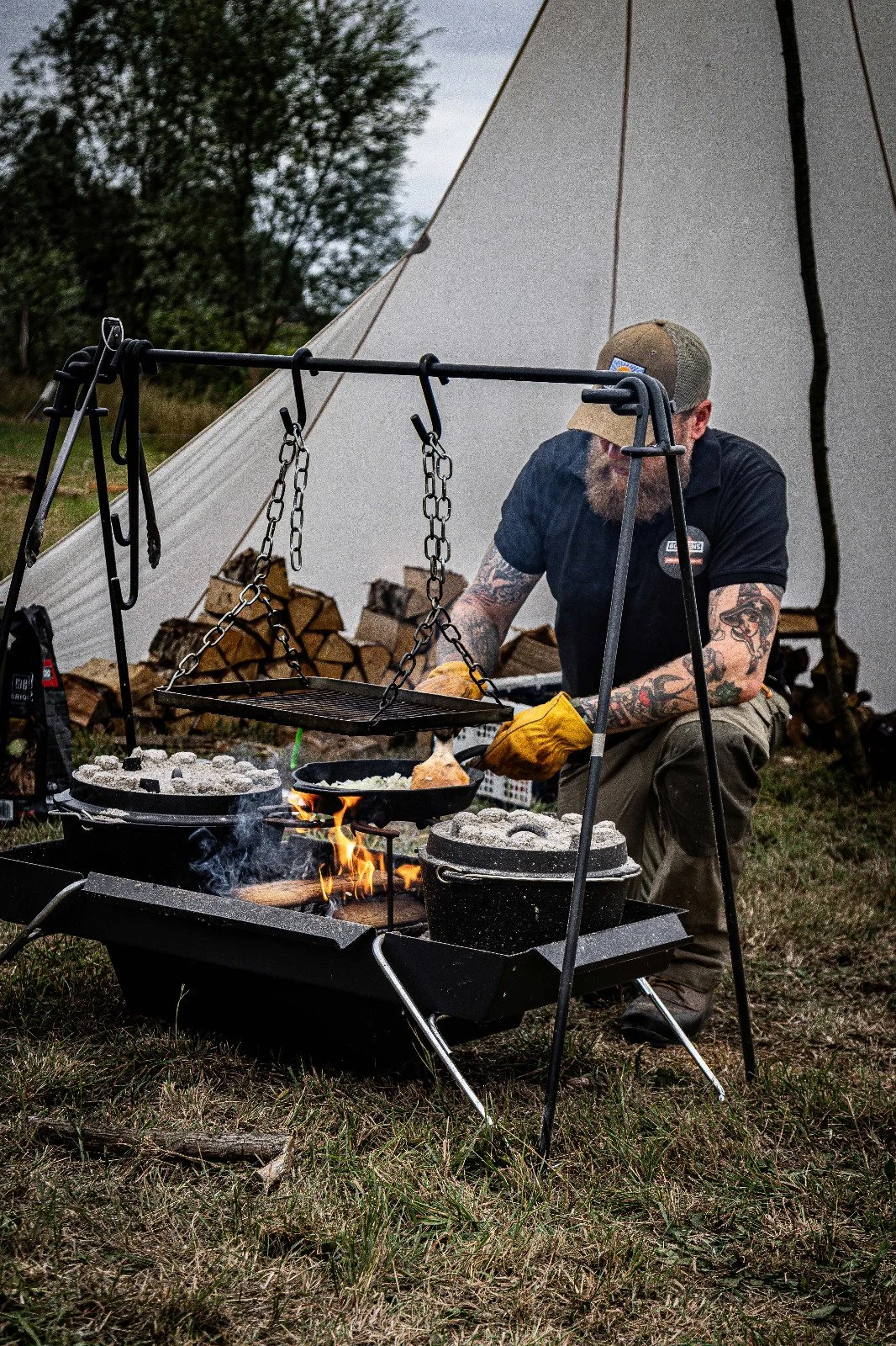 Man with tattoos cooking outdoors over an open flame, grilling chicken and vegetables, with a camping tent and stacked firewood in the background.