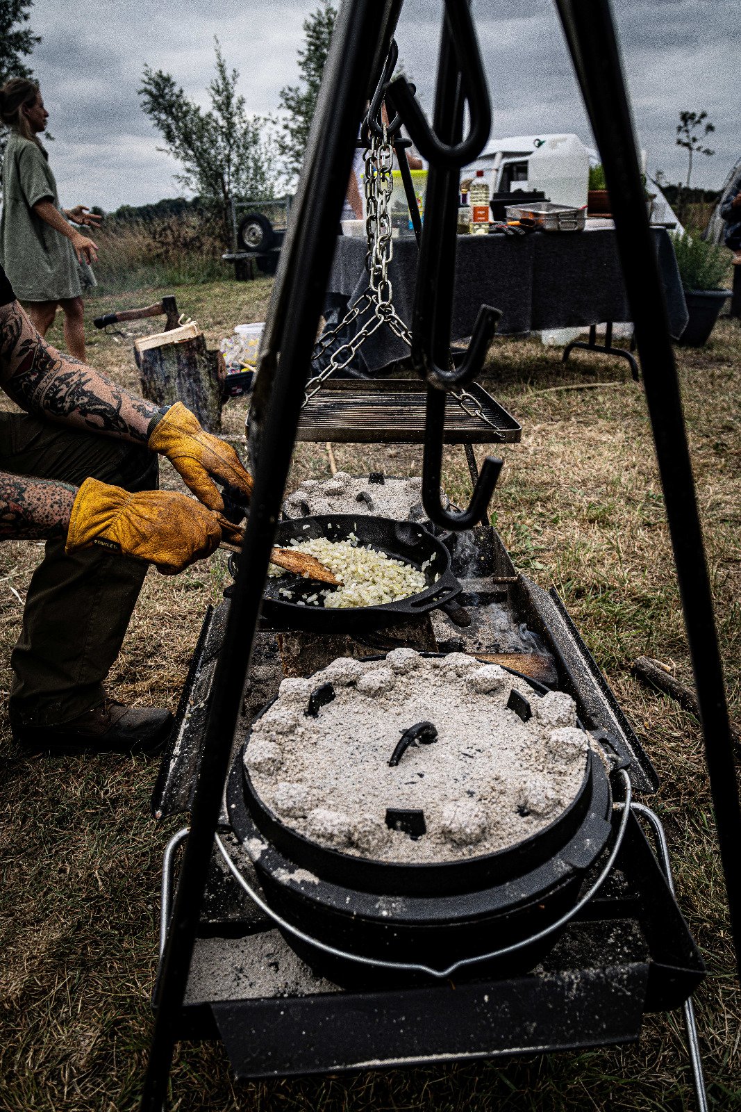 Person cooking onions over an outdoor fire on a cast iron skillet, with a grill covered in salt or flour in the foreground, set at an outdoor gathering.