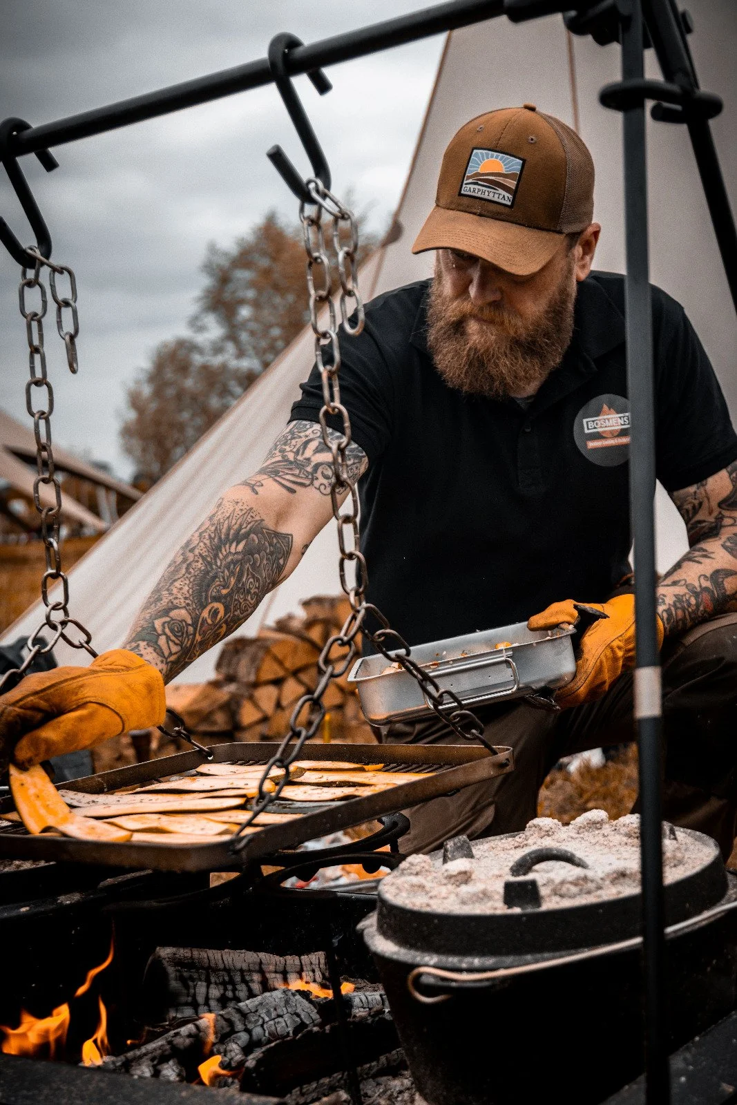 A man with tattoos, a beard, and wearing a cap and gloves is grilling meat over an open fire outdoors, with a backdrop of wood logs and a tent.