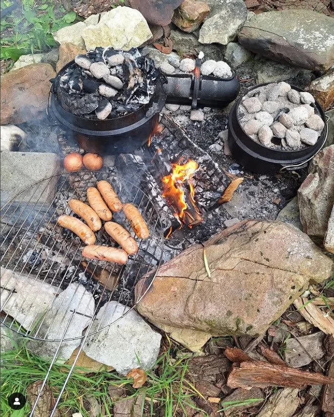 A cooking setup outdoors with two black cookers filled with rocks and charcoal, a small open fire burning in the center, and a wire rack with sausages grilling over the fire. There are also two eggs on the side. The area is surrounded by rocks, wood,