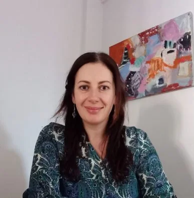 A woman with long dark hair smiling in front of a colorful abstract painting on a white wall.