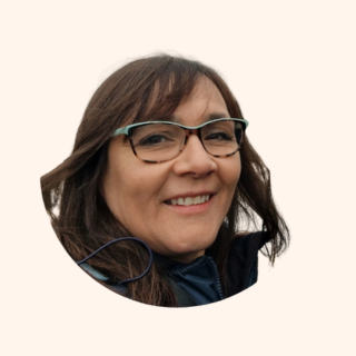 Smiling woman with shoulder-length brown hair and glasses, wearing a dark top, against a plain background.
