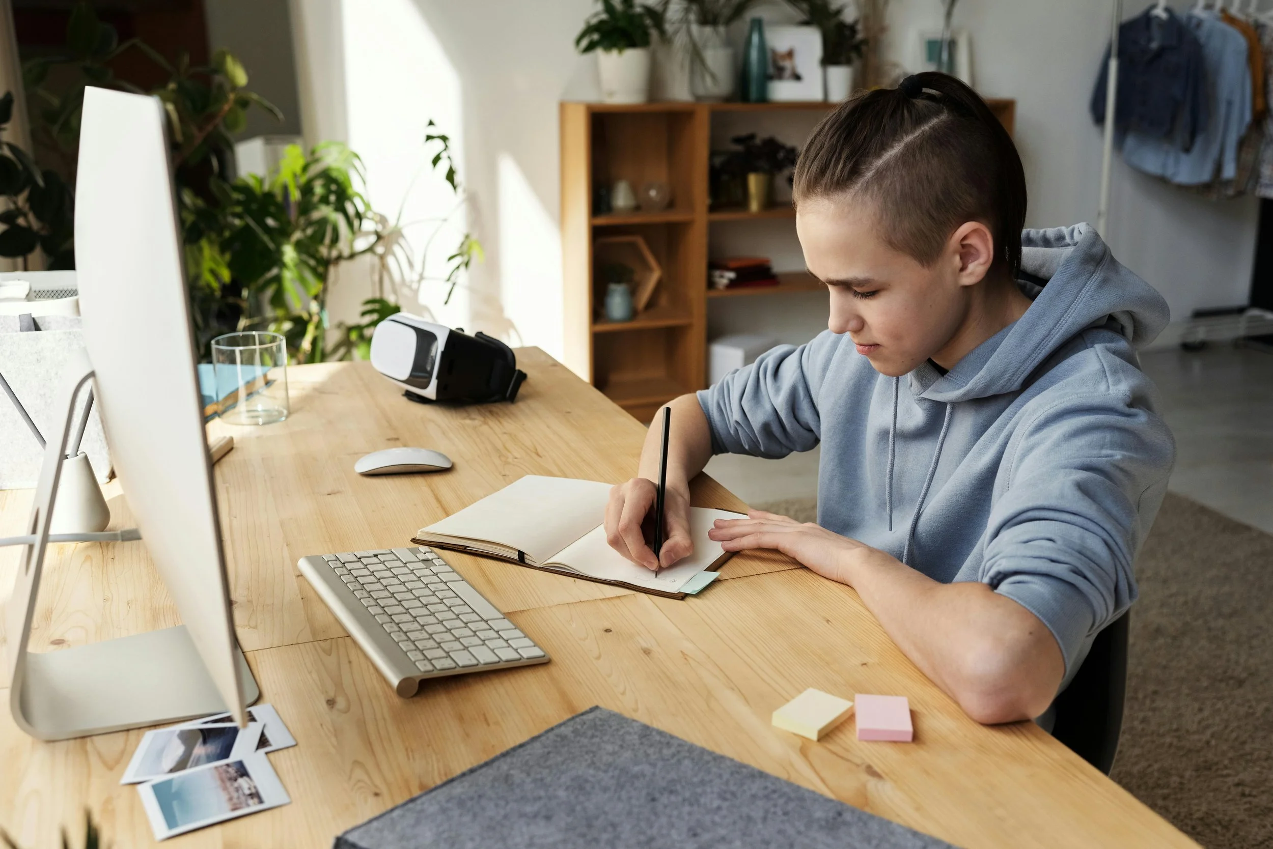 Person with short hair writing in a notebook at a wooden desk, with computer, keyboard, mouse, and VR headset, in a well-lit room with plants and shelves.