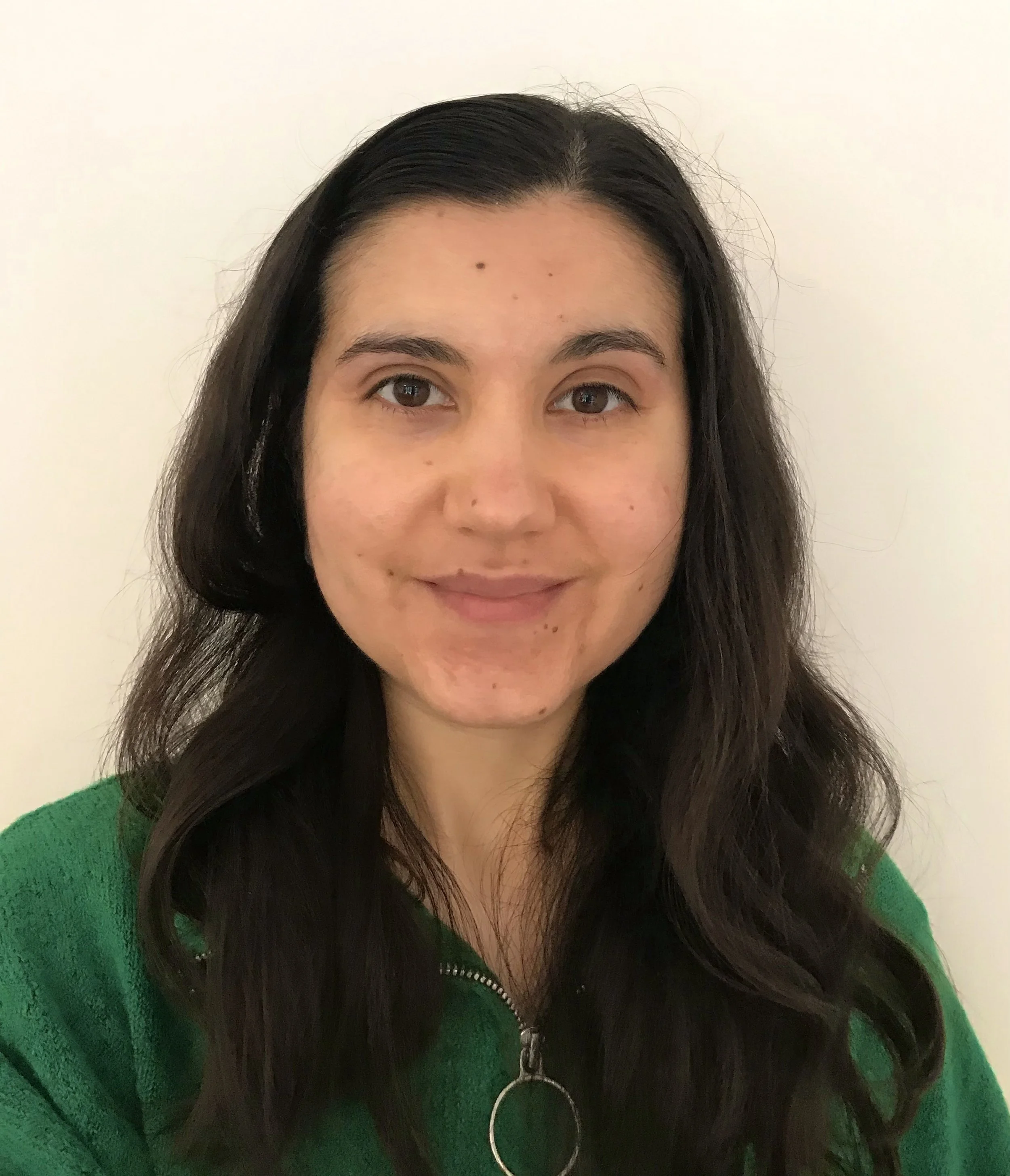 Portrait of a woman with long dark hair, wearing a green zip-up sweater, standing against a plain light-colored background.