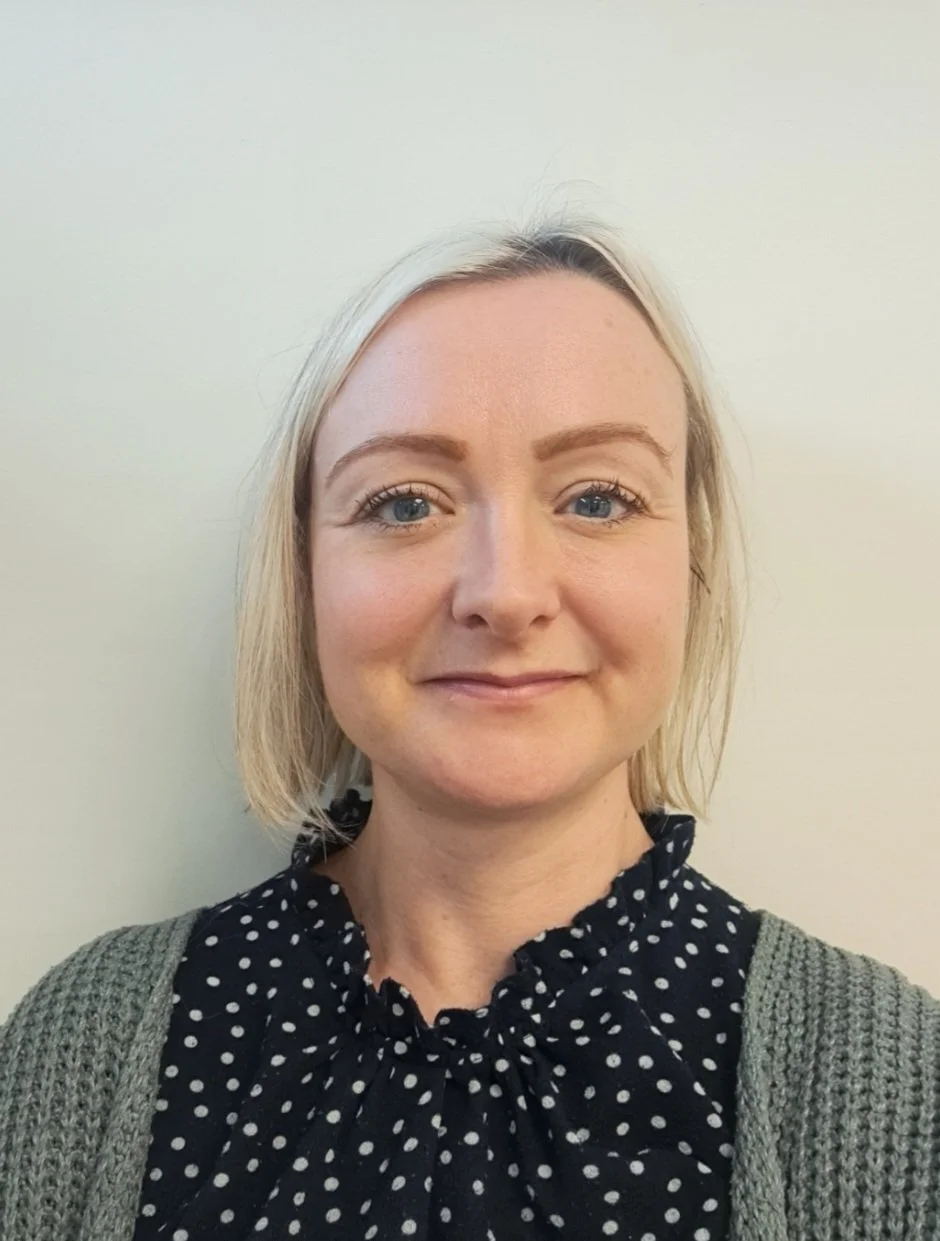 A woman with blonde hair, blue eyes, and fair skin, smiling, wearing a black polka dot blouse and gray cardigan, against a plain pale background.