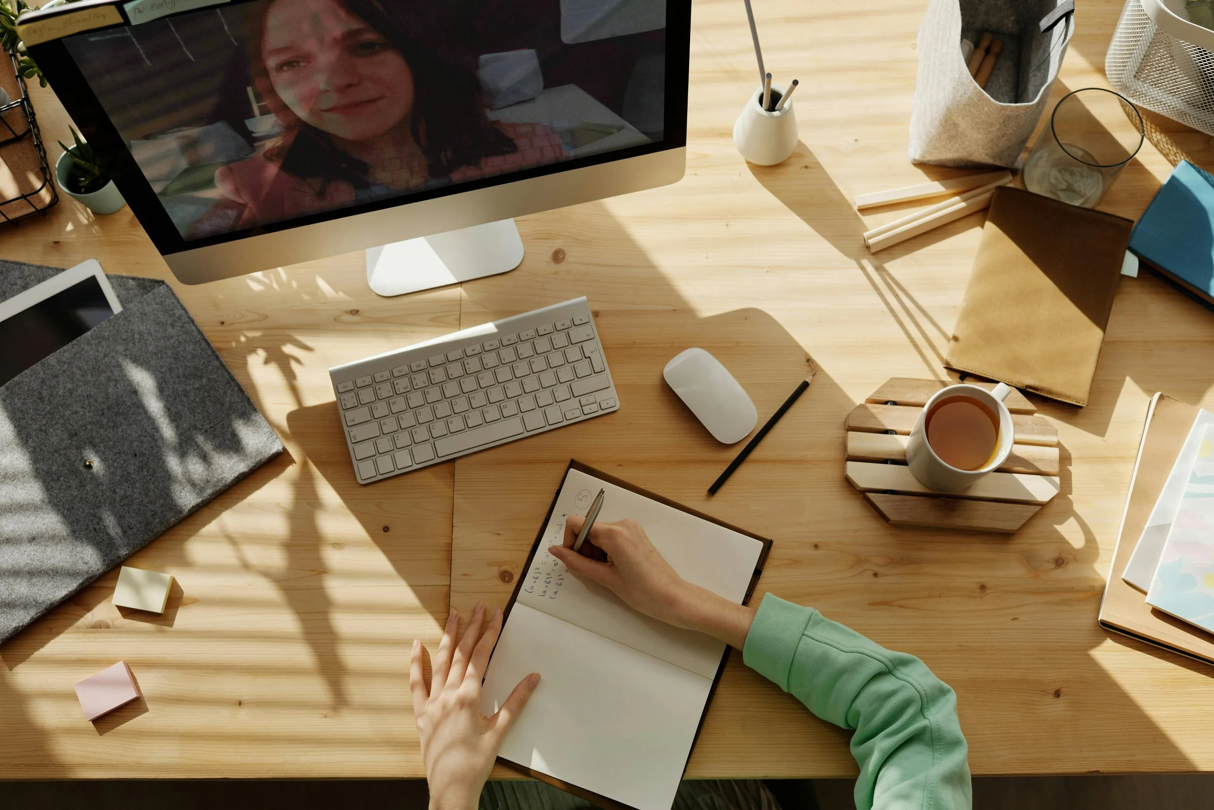 A person working at a wooden desk with a notebook, pen, and a cup of tea, while attending a video call on an iMac computer. The desk also has a wireless keyboard and mouse, a small plant, stationery, notebooks, and various office supplies.