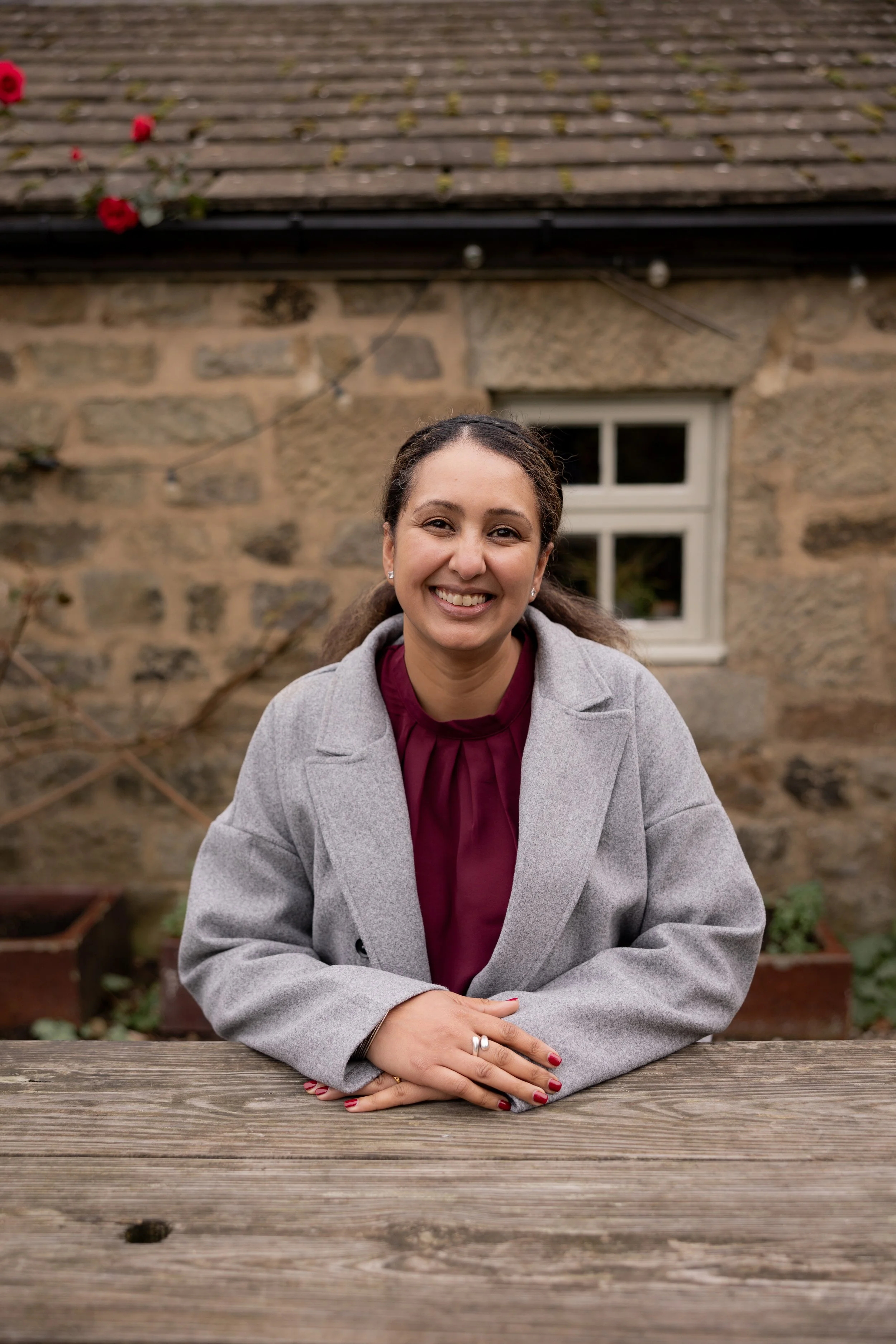 Smiling woman sitting at a wooden table outside, wearing a gray coat and a burgundy blouse, with a stone house and window in the background.