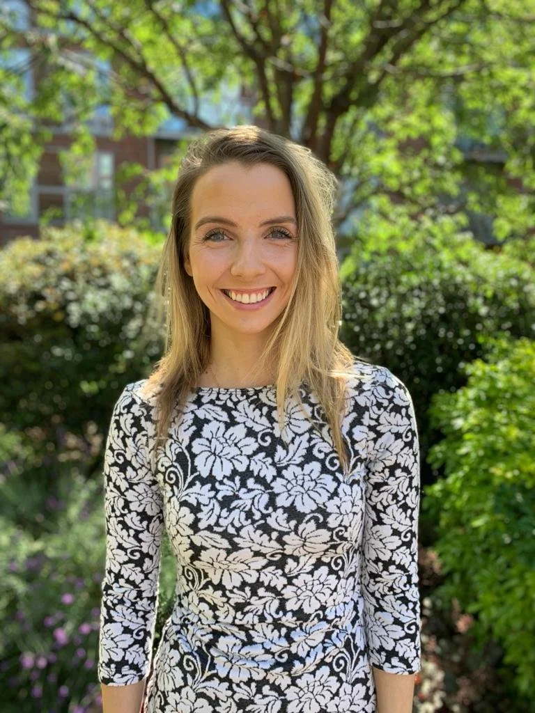 A young woman standing outdoors in a garden, smiling at the camera, wearing a black and white floral dress with greenery and trees in the background.