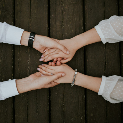 Two pairs of hands holding each other over a wooden surface, one pair with a wedding ring and the other with a bracelet.