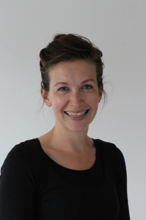 A woman with brown hair styled in an updo, smiling, wearing a black top against a plain light background.