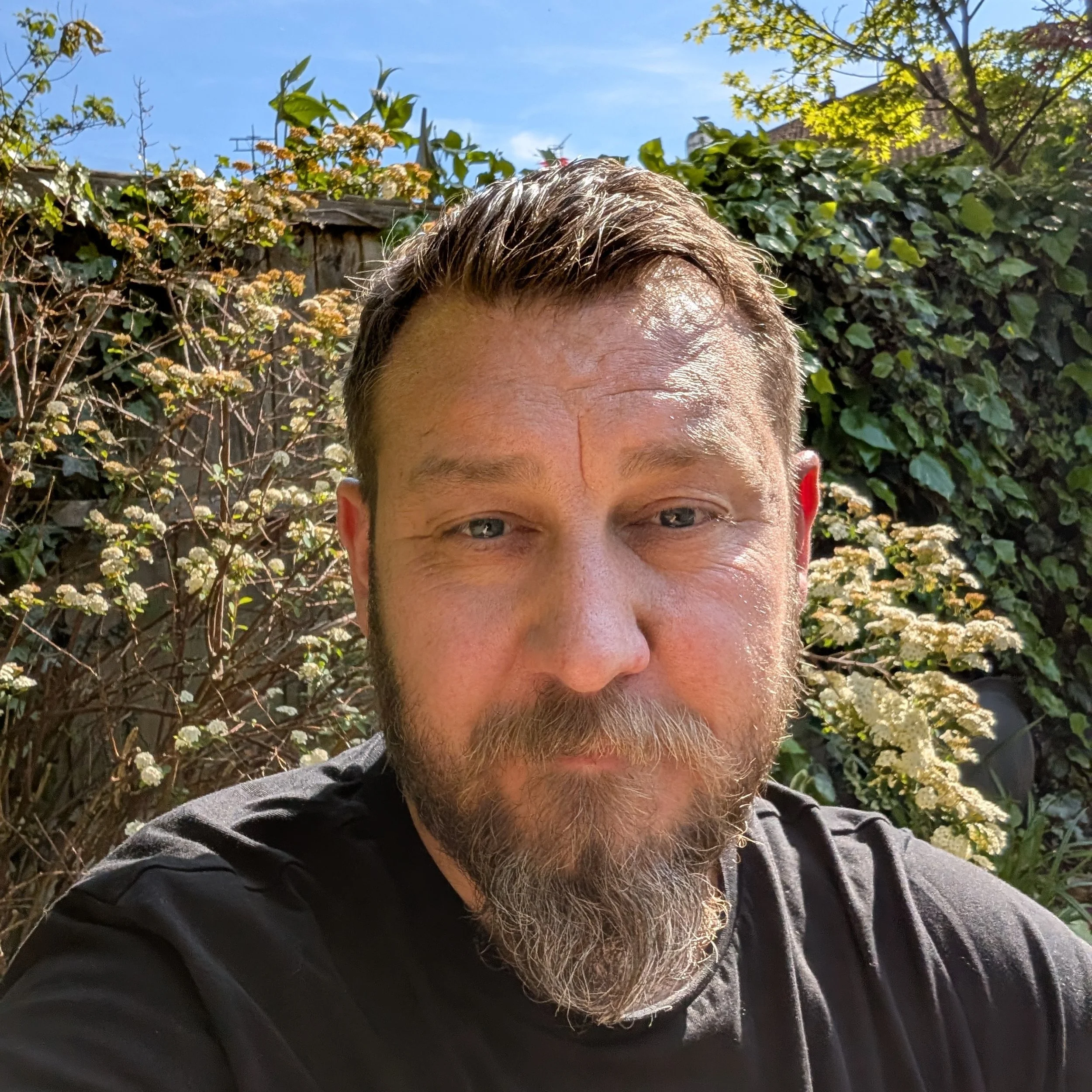 A man with short brown hair, a beard, and light skin takes a close-up selfie outdoors in a garden with green plants and white flowers, under a clear blue sky.
