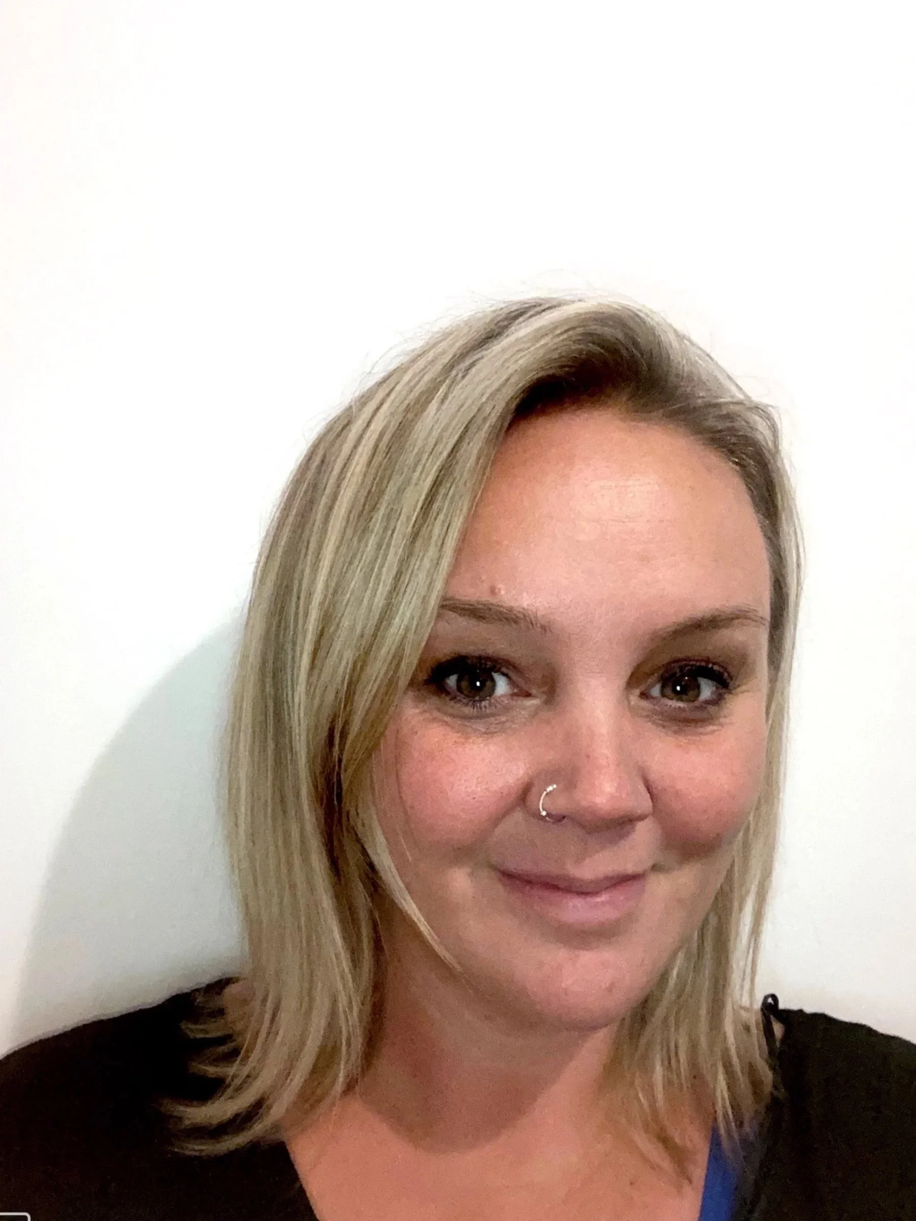 A woman with shoulder-length blonde hair, brown eyes, and a nose piercing, smiling with a white wall in the background.