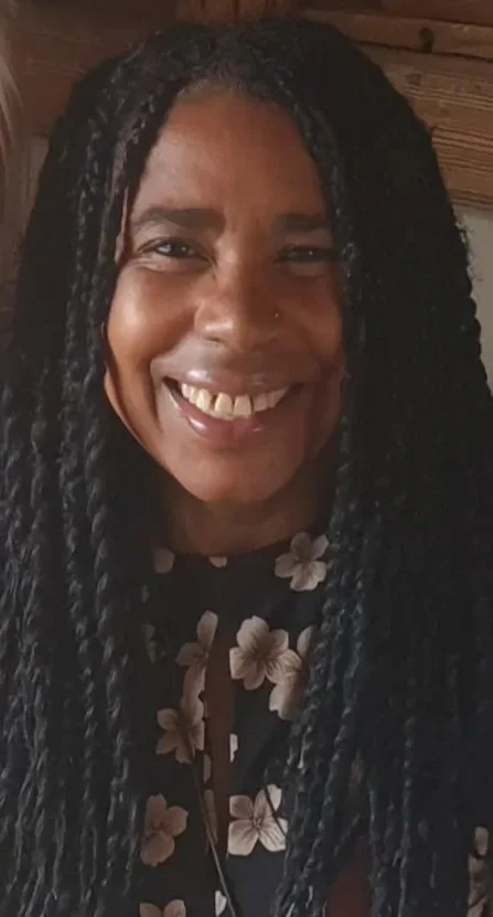 A woman with long black twisted hair smiling at the camera, wearing a floral top with a wooden background.