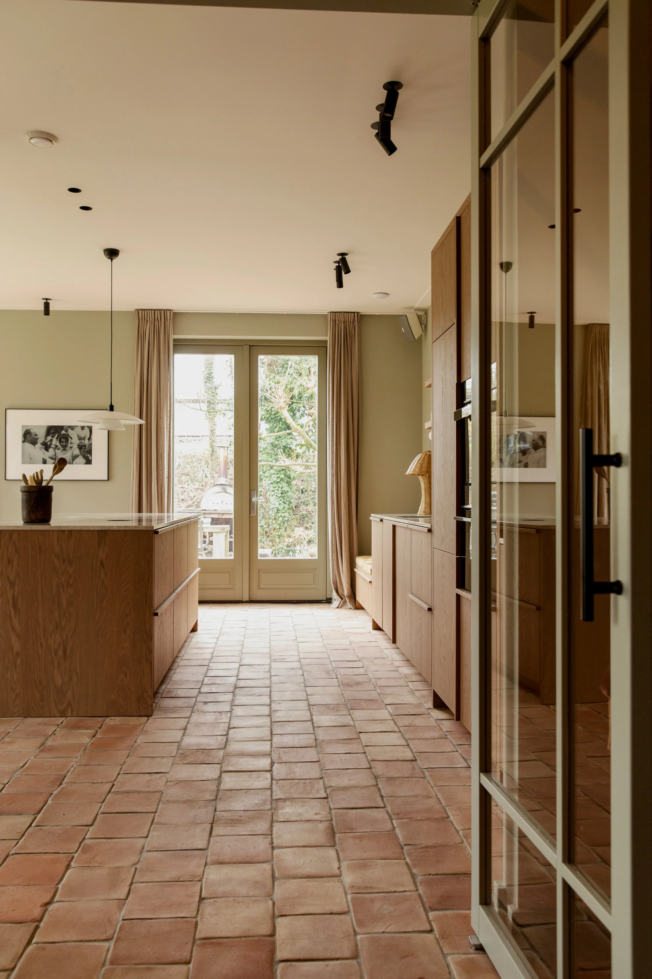 Modern kitchen with terracotta tile floor, wooden cabinetry, glass door leading outside, beige curtains, and black ceiling lights.