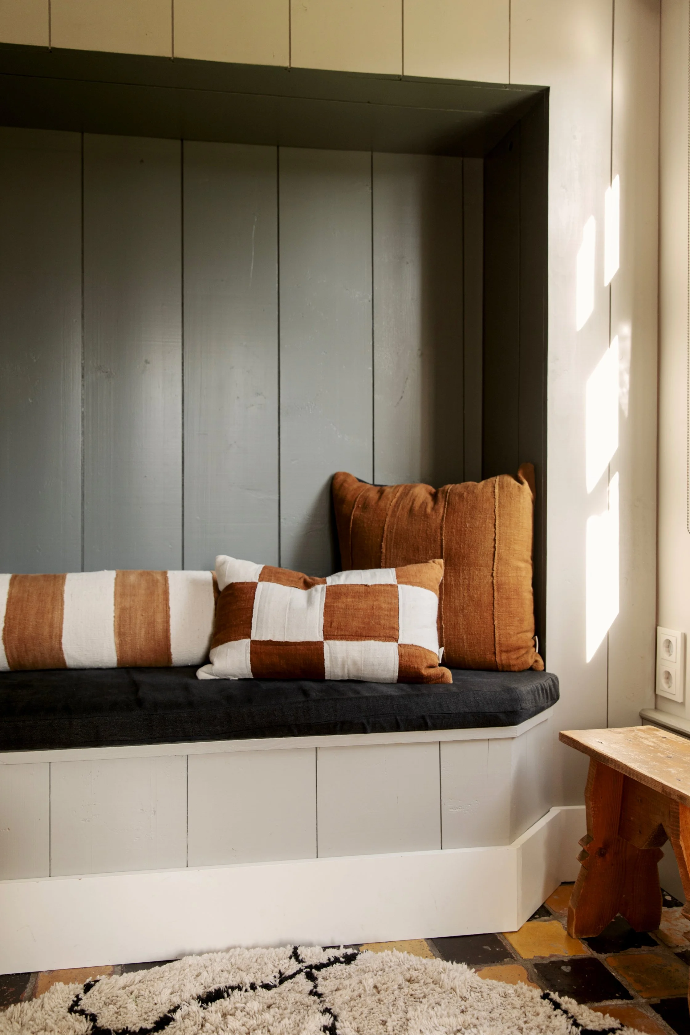A cozy built-in window seat with black cushion and rust-colored pillows, next to a window with sunlight, a wooden side table, and a patterned rug on tiled floor.