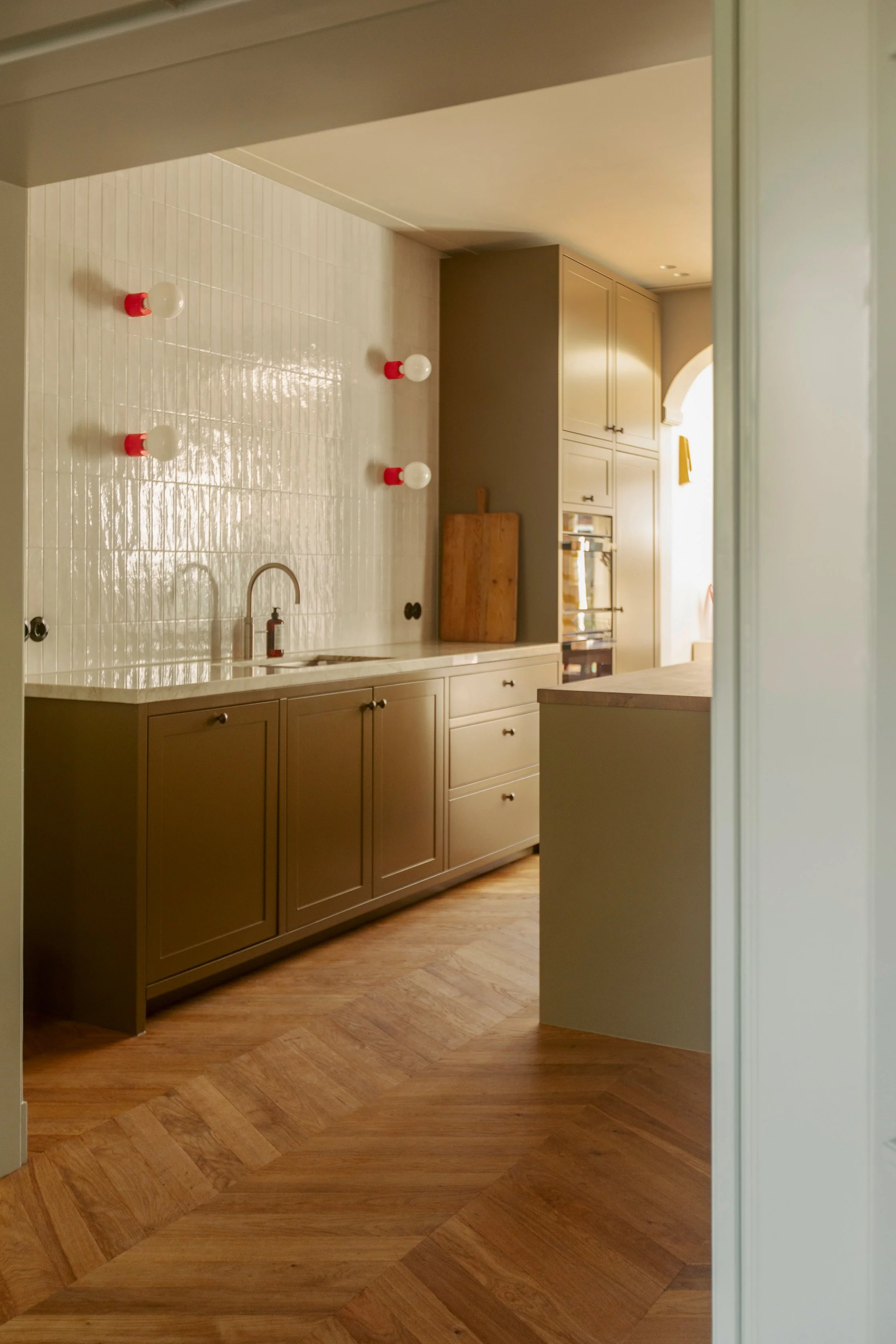 Empty kitchen with beige cabinets, a white tiled backsplash, and four light bulbs on a wall. Wooden cutting board near a doorway with bright light coming in.