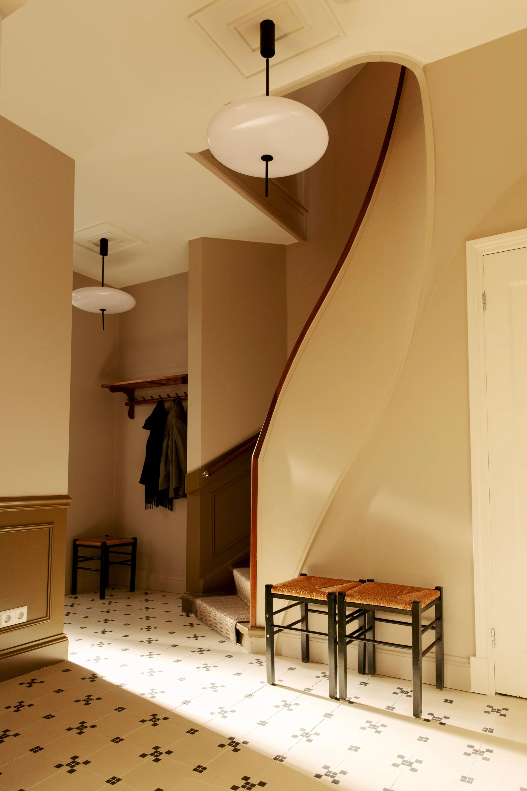 Interior entryway with a staircase, two hanging ceiling lights, two chairs with woven seats, and a coat rack with jackets.