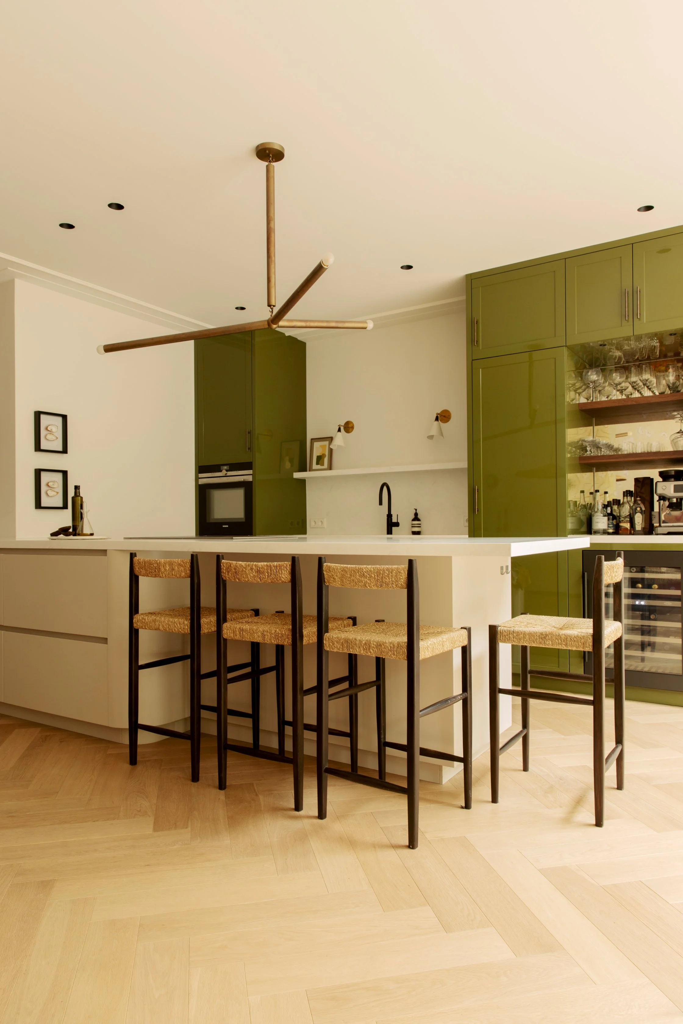 Modern kitchen with a white island counter, four high wooden chairs with woven seats, green cabinets, a built-in oven, and open shelves with glassware and bottles.