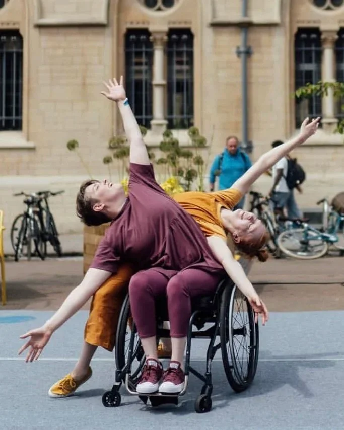 2 performers outside in the public square.  One performer is seated in a wheelchair, while the other leans across her; their bodies intertwined. Both extend their arms wide, and gaze upwards, creating a sense of openness, trust and connection.