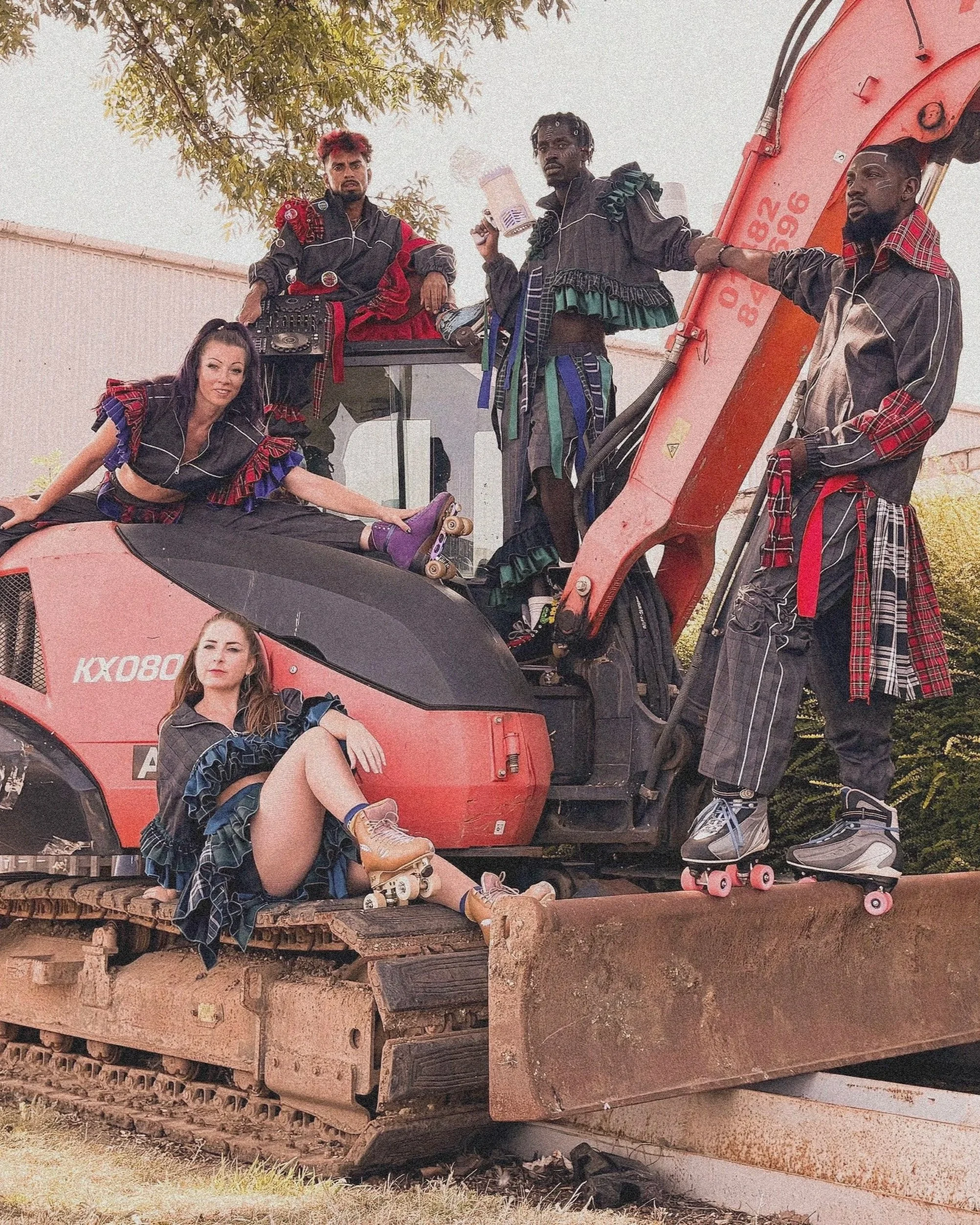 5 performers dressed in matching avante-guarde style jump suits posed around a tractor outdoors wearing rollerskates.