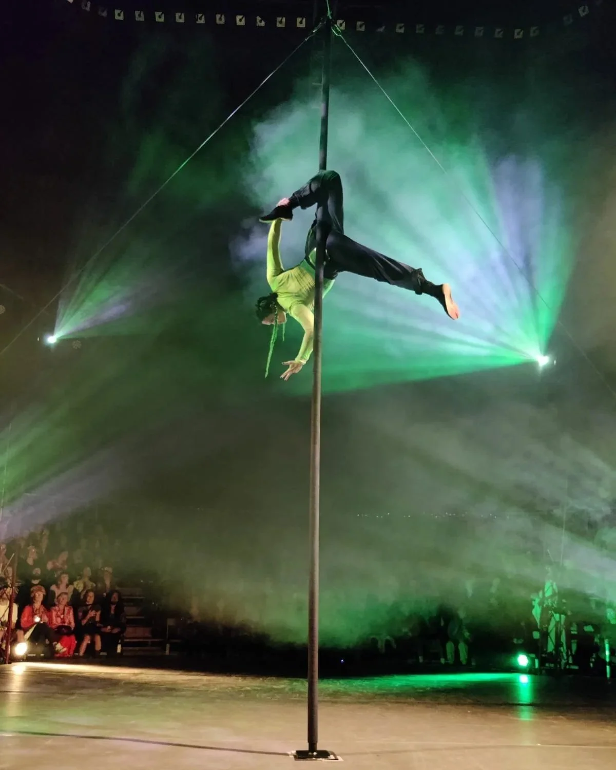 A circus performer balances on a large vertical pole at the centre of a circus tent. Suspended several metres above the ground, she is holding a striking pose with one leg hooked around the pole and the other extended outward, while one arm reaches.