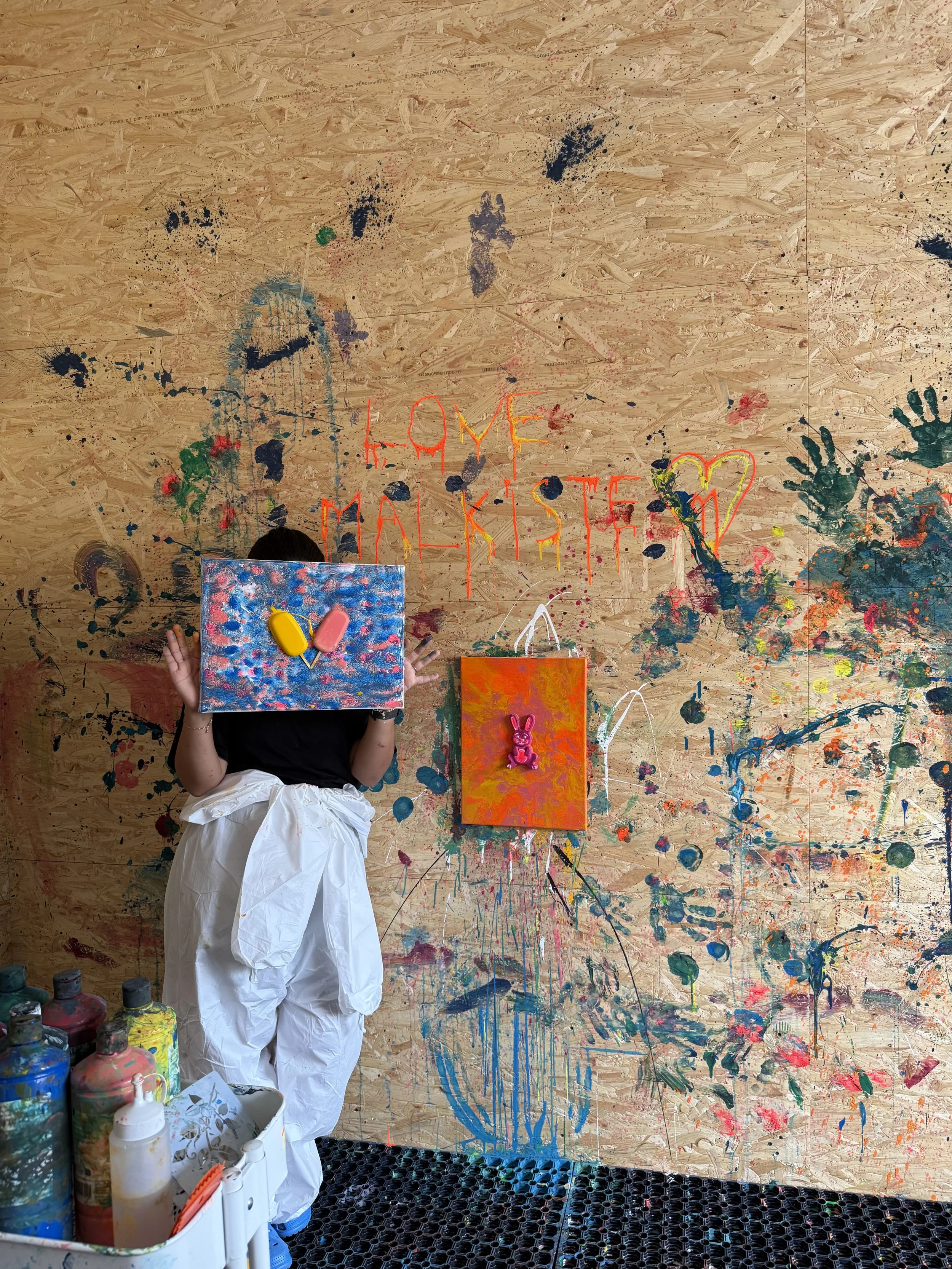 A child wearing a white paint smock, standing in front of a large wooden wall covered with colorful paint splatters and handprints. The child is holding two painted canvases, one with blue and white colors, and the other orange with a pink bunny sticker. The wall has the words 'Home Maker' written in red and yellow paint.