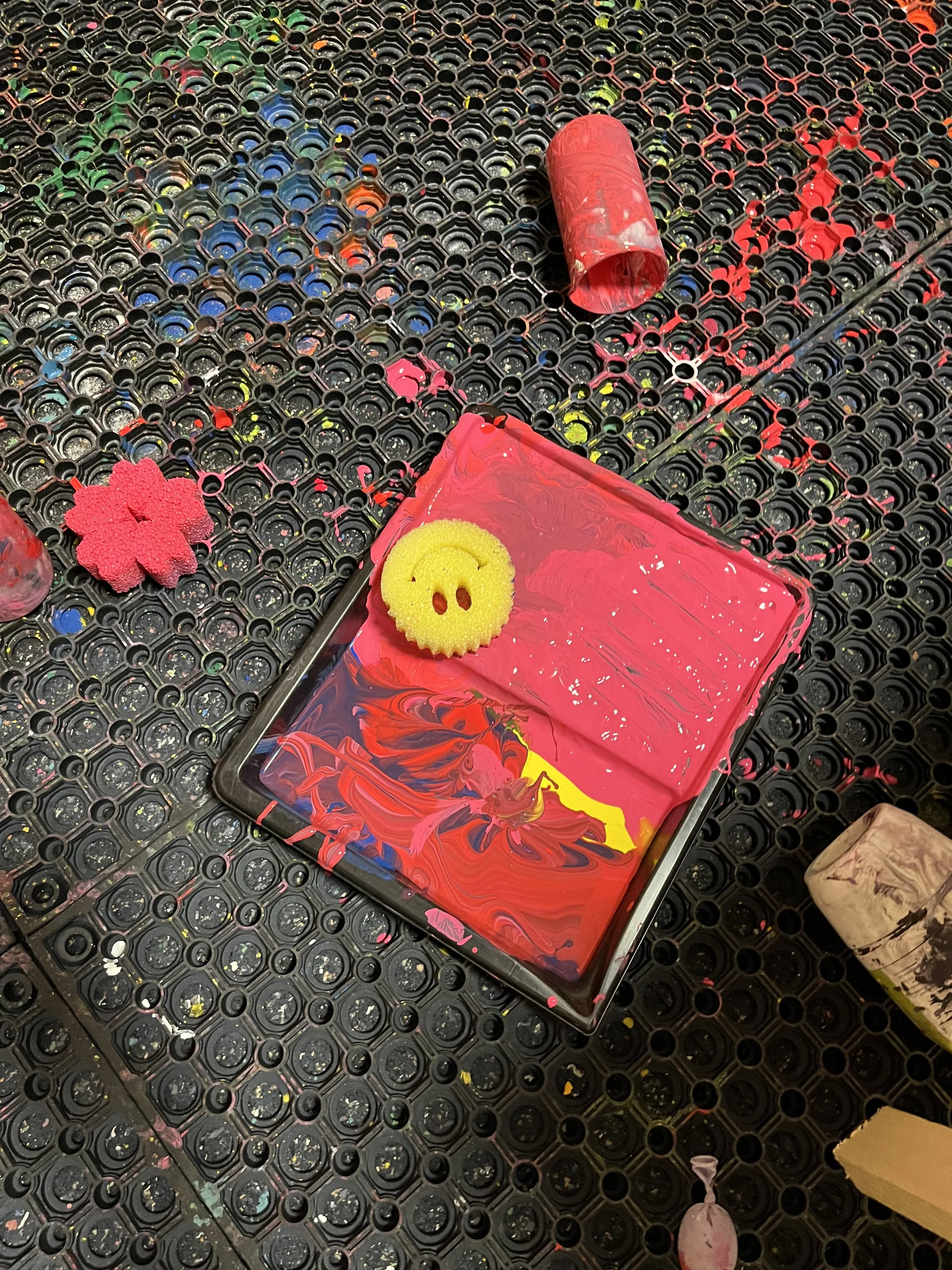 A black rubber floor mat with colorful paint splatters, a red paint roller, a sponge with a smiley face, a pink sponge in the shape of a flower, and a cardboard tube.