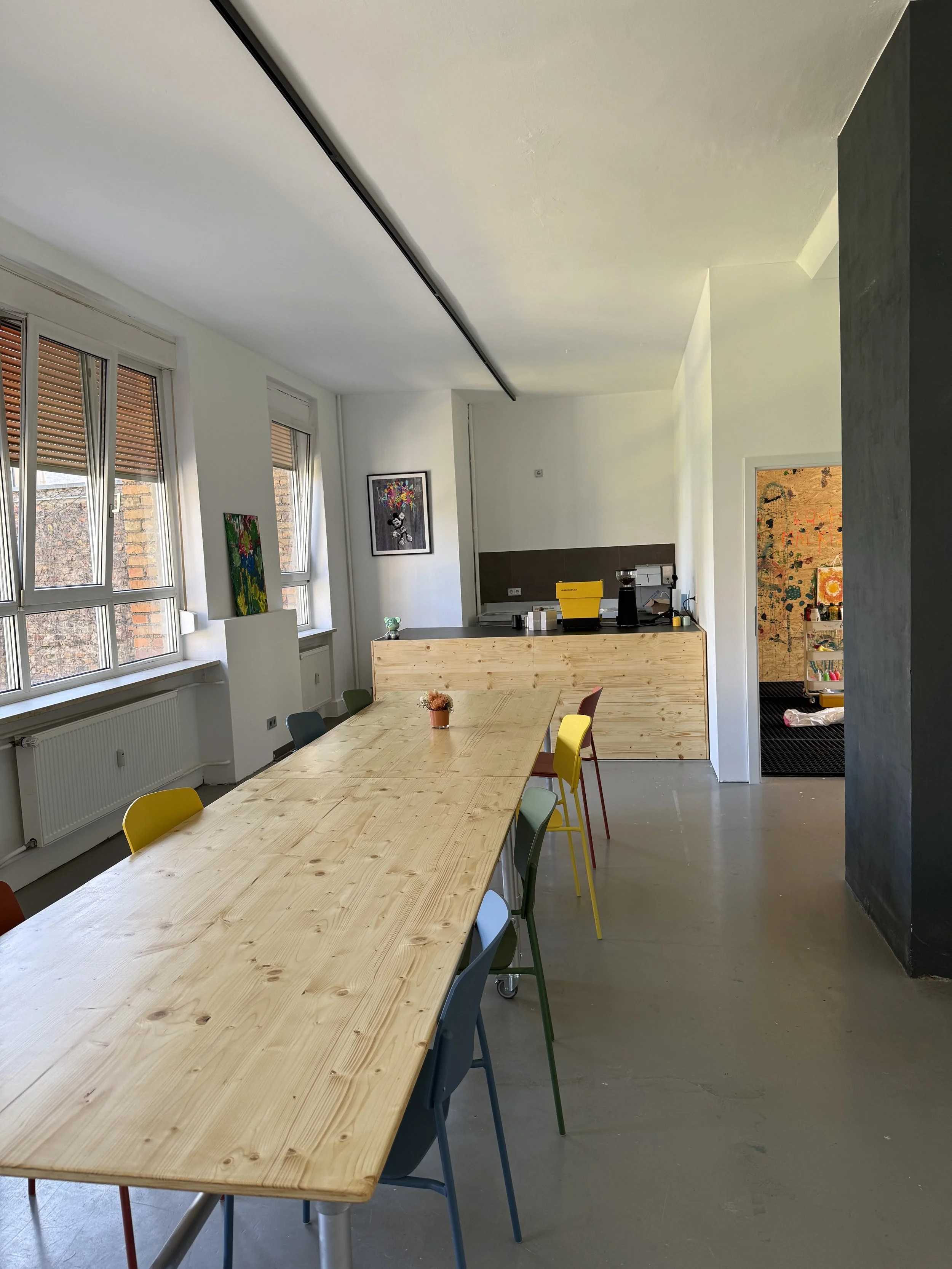 A conference room or classroom with a long wooden table and colorful chairs. Large windows with blinds, artwork on the walls, and a small snack bar or coffee station in the background.