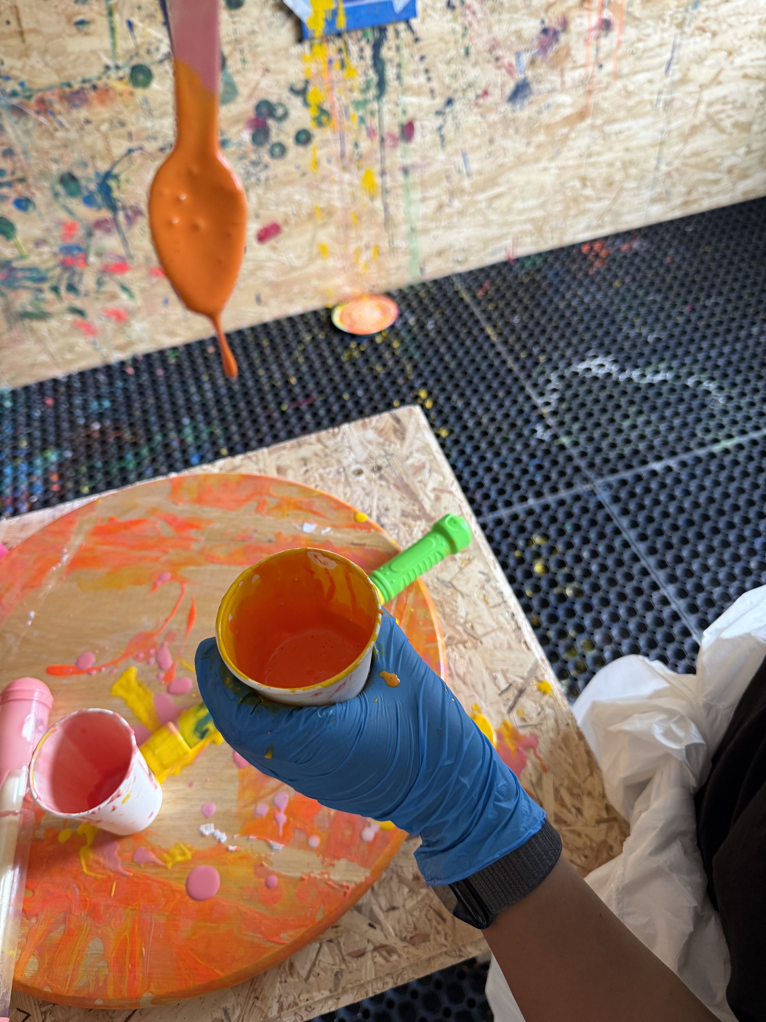 Person wearing a blue glove holding a cup of orange paint in an art studio with scattered paint and a paint-covered table.
