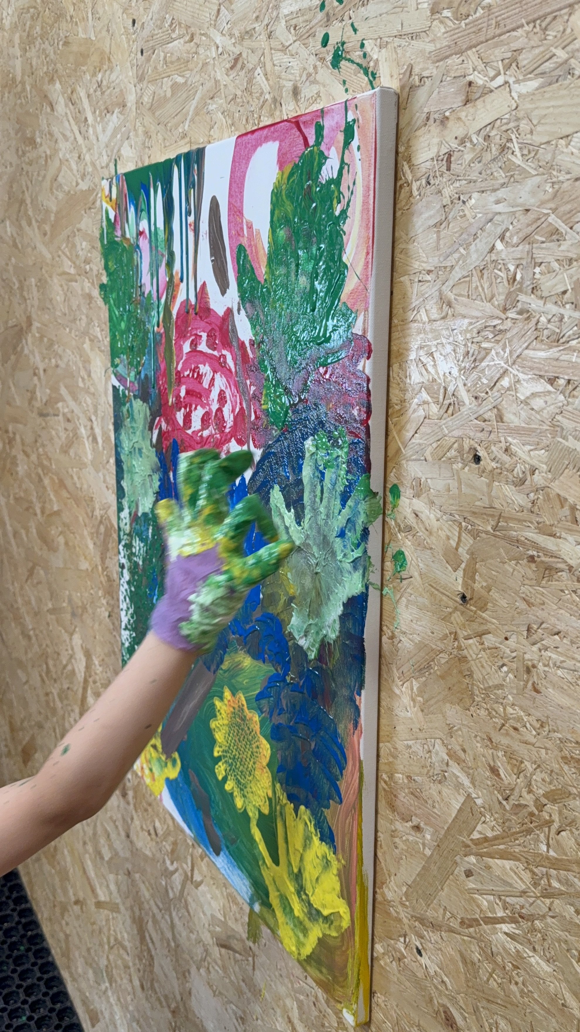 Child's hand with purple glove painting on a large canvas with colorful acrylic paints, depicting sunflowers and a lush landscape.