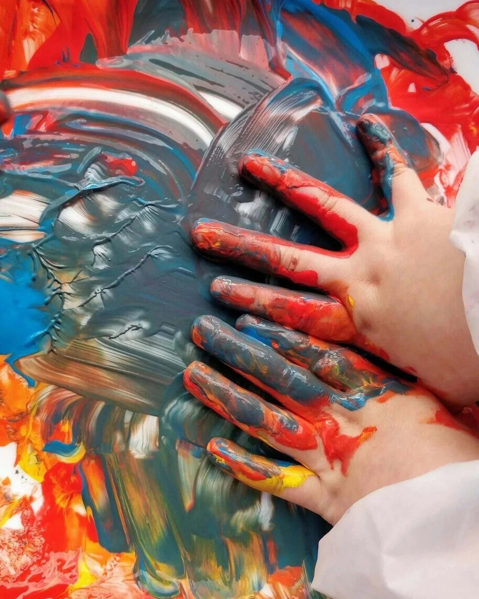Close-up of a hand covered in colorful paint, pressing against a surface covered in swirling, vibrant red, blue, yellow, orange, green, and white paint.