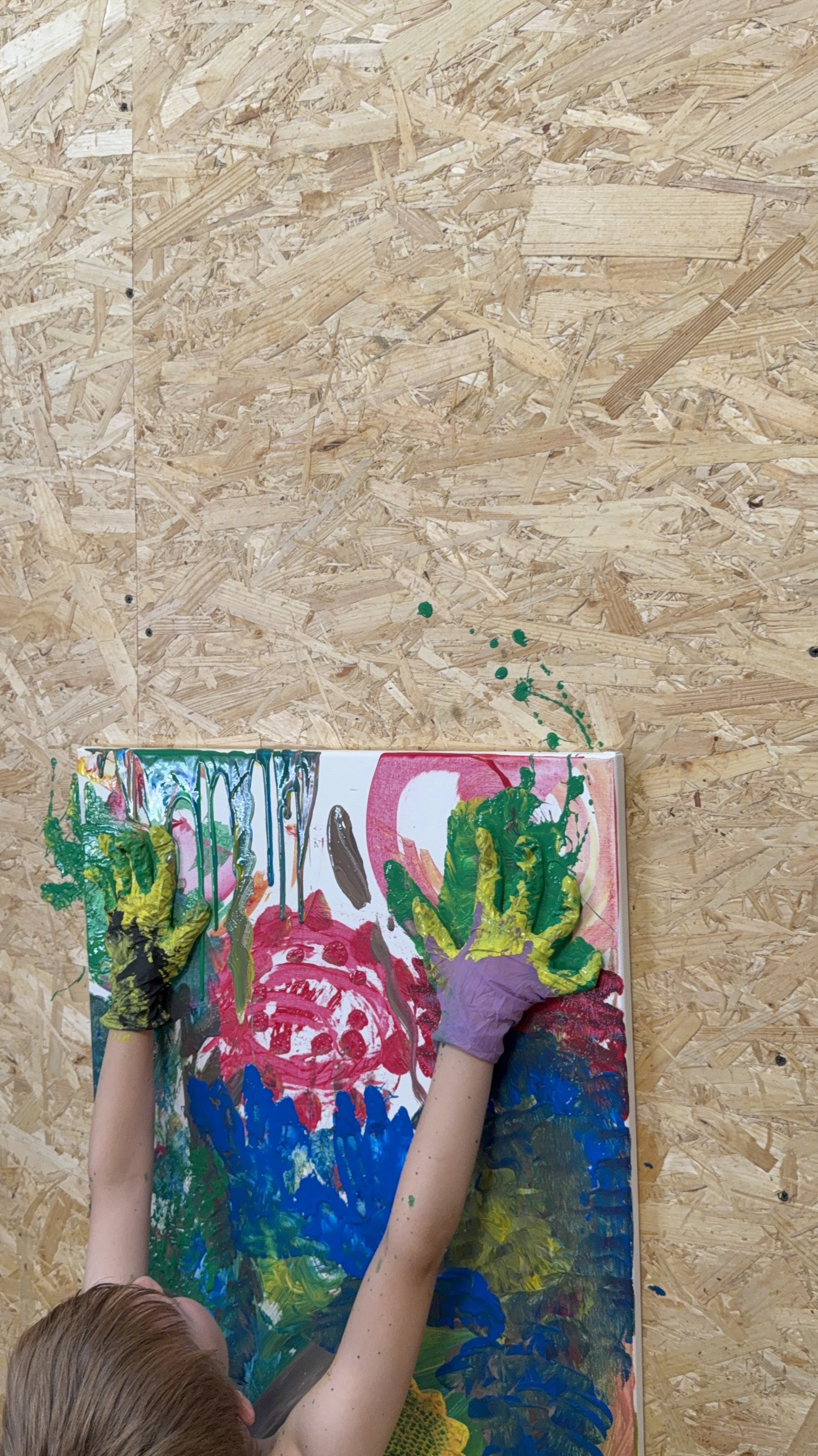 Child's hands covered with colorful paint, working on a vibrant abstract painting on a canvas on a wooden surface, with green paint splatters on the background.