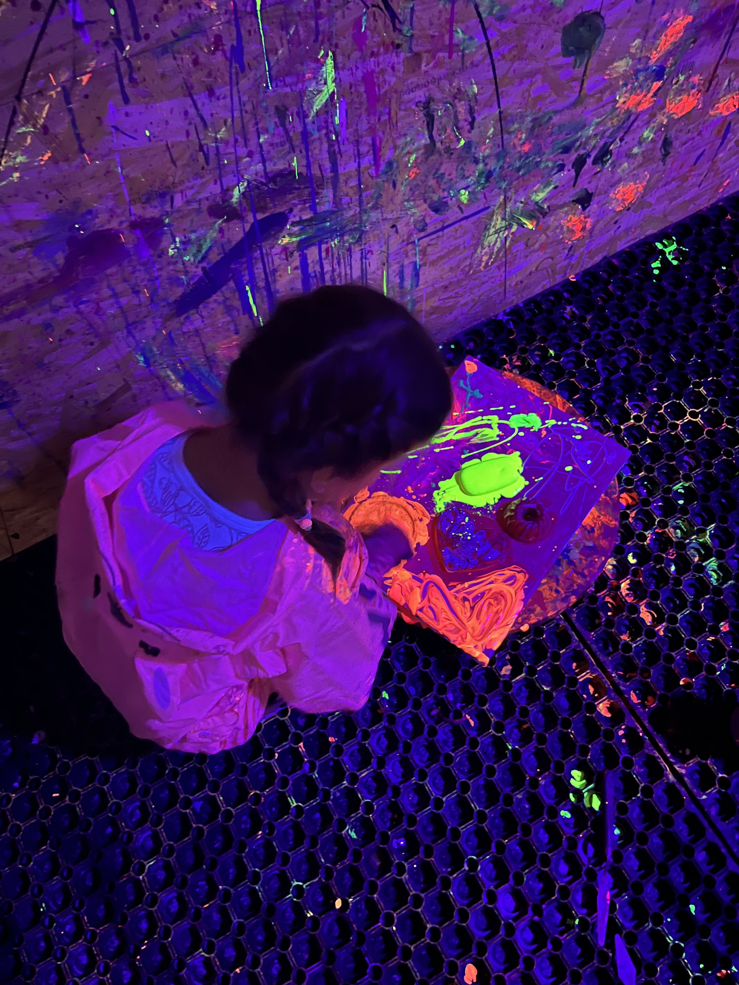 A young girl with dark hair in braids wearing a white dress, sitting on a black honeycomb-patterned floor, illuminated by blacklight. She is drawing glowing neon images of cartoon characters and shapes on a table in front of her, which is covered wit