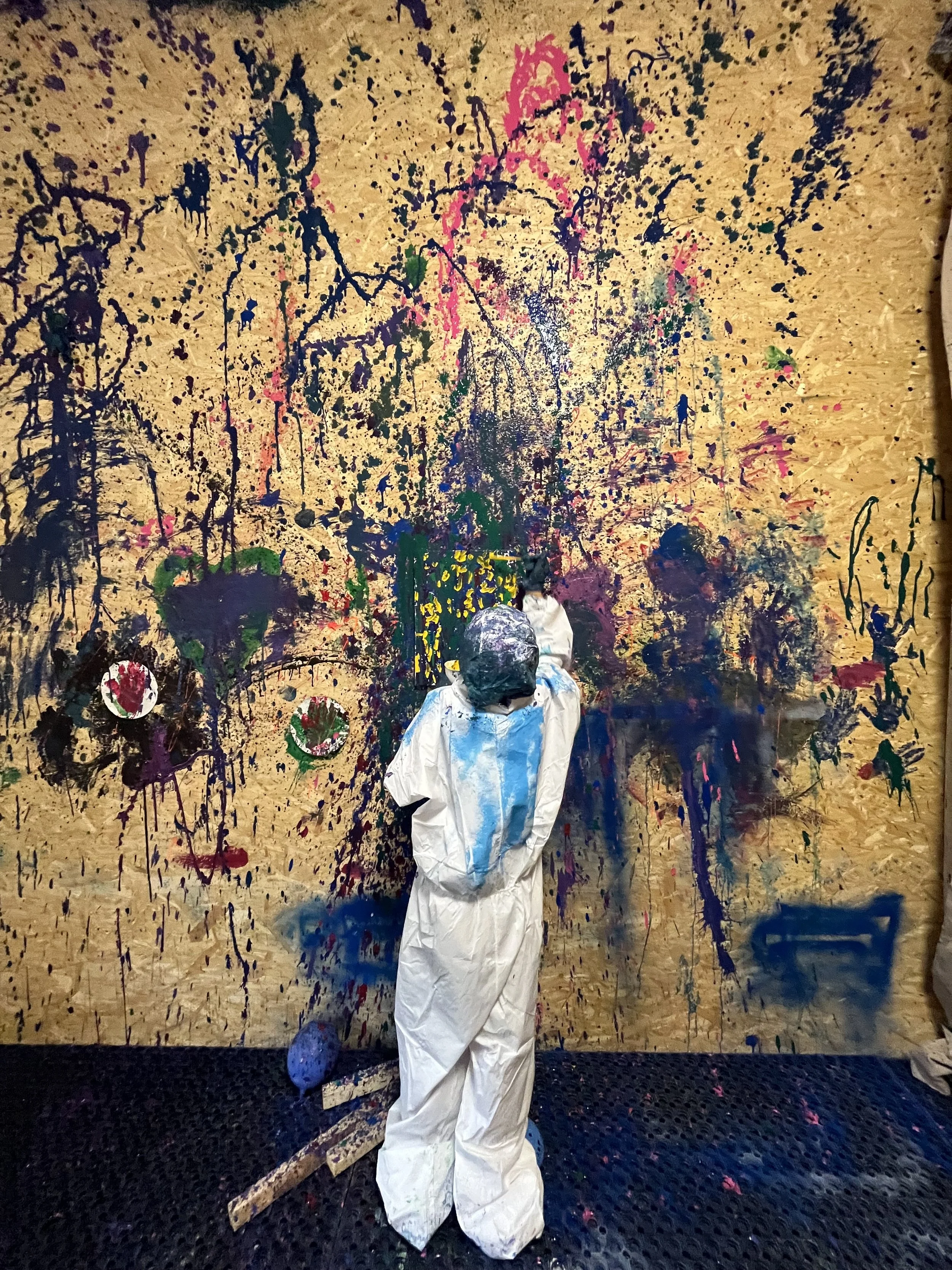 Child dressed in a white protective suit with paint-stained shoes, standing in front of a large, colorful abstract painting on a wooden wall, created with splatters and drips of various paint colors.