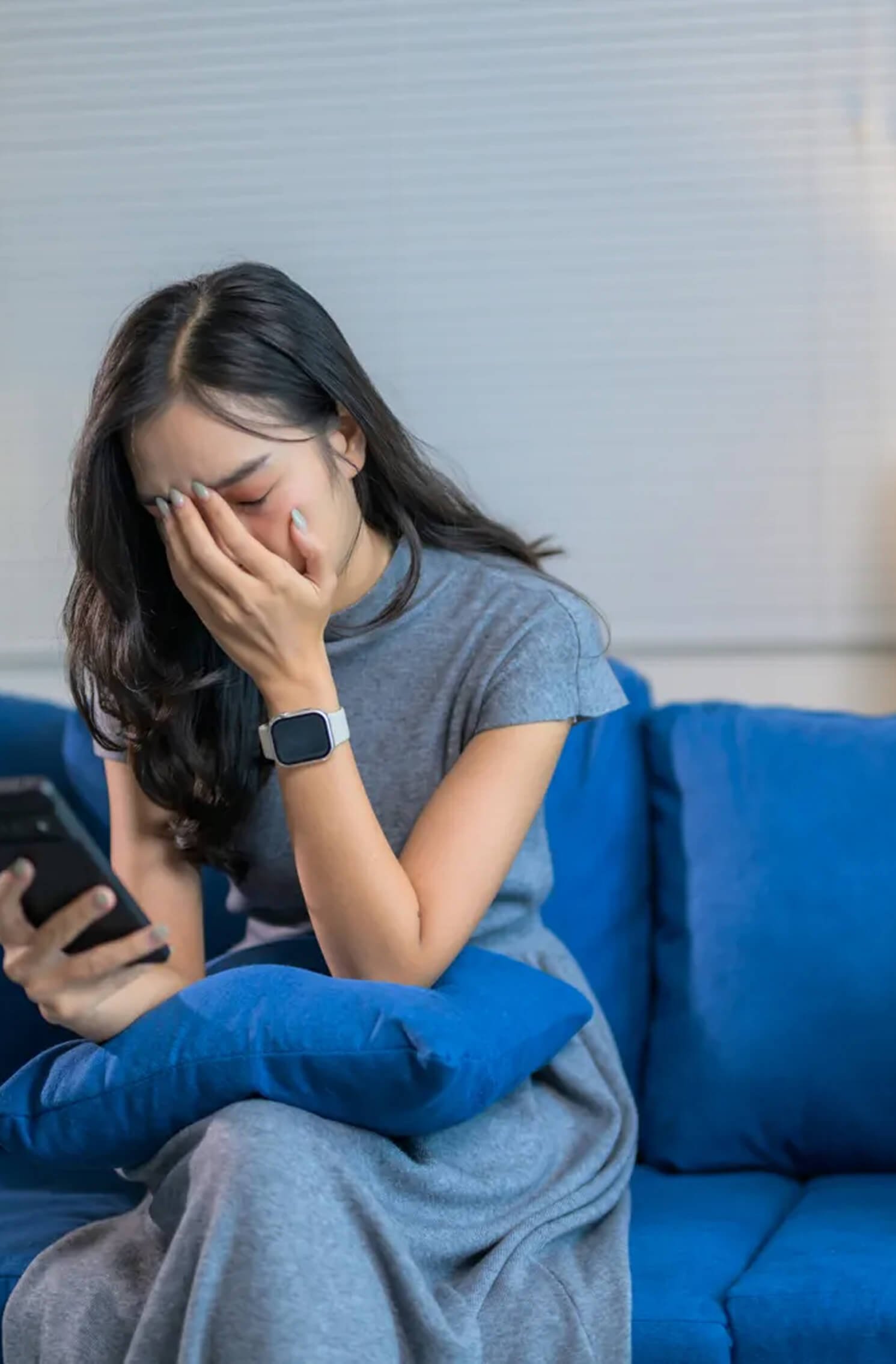 A woman sitting on a blue couch, holding her face with one hand and looking at her phone with a worried expression.