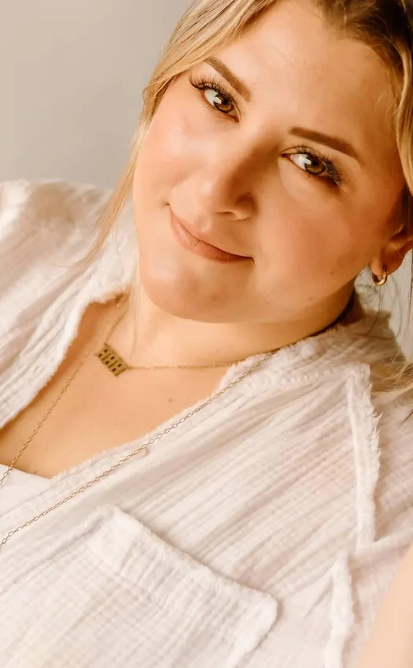 A woman with blonde hair, brown eyes, and gold jewelry, smiling gently at the camera, wearing a cream-colored textured jacket.