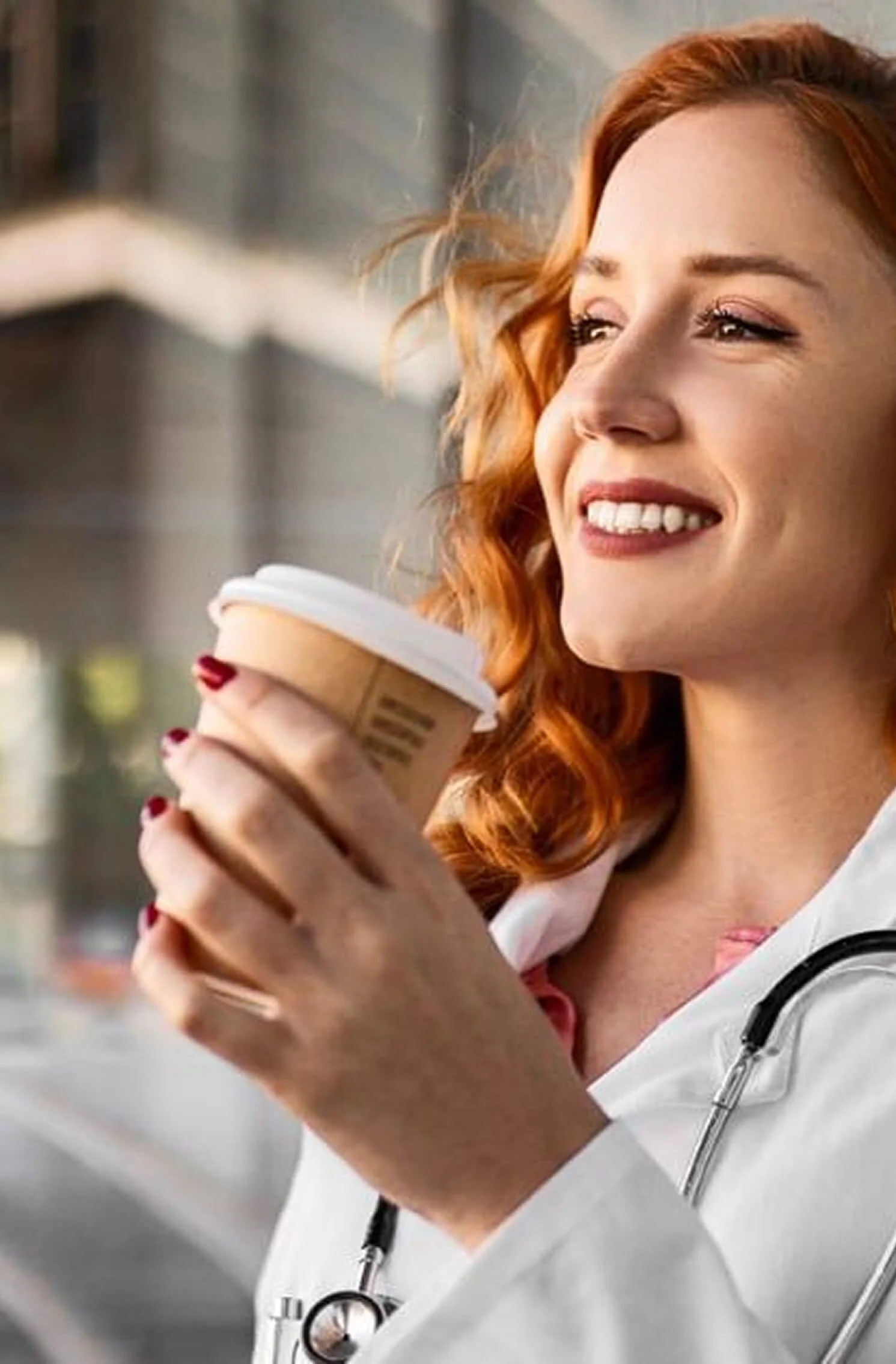 A woman with red hair, wearing a white medical coat and a stethoscope, holding a to-go coffee cup and smiling.