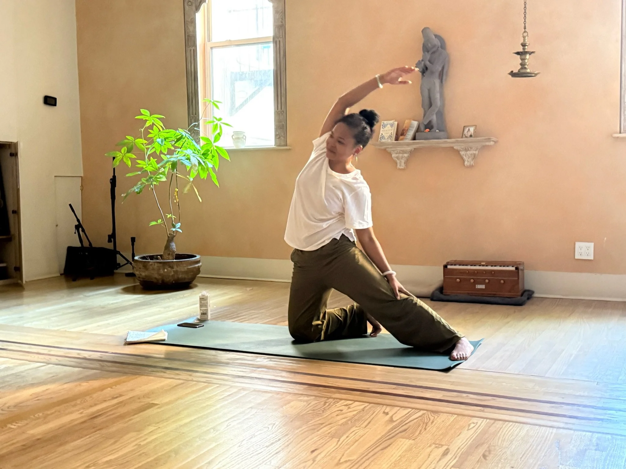 A person practicing yoga in a room with beige walls and hardwood floors. The person is on a yoga mat, performing a pose with one arm extended overhead and the other arm reaching behind, holding their ankle. There is a large green plant on the left, a window with natural light, a fireplace mantle on the right with picture frames and a sculpture, and a small wooden box on the floor.