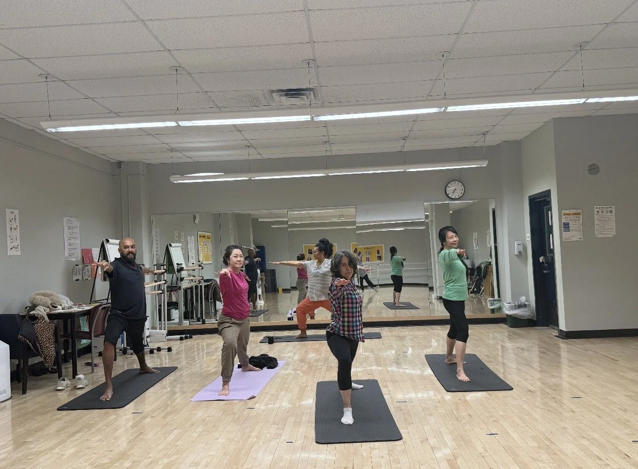 Group of people participating in a yoga or fitness class in a studio, standing on mats and performing a yoga pose with one arm extended out.