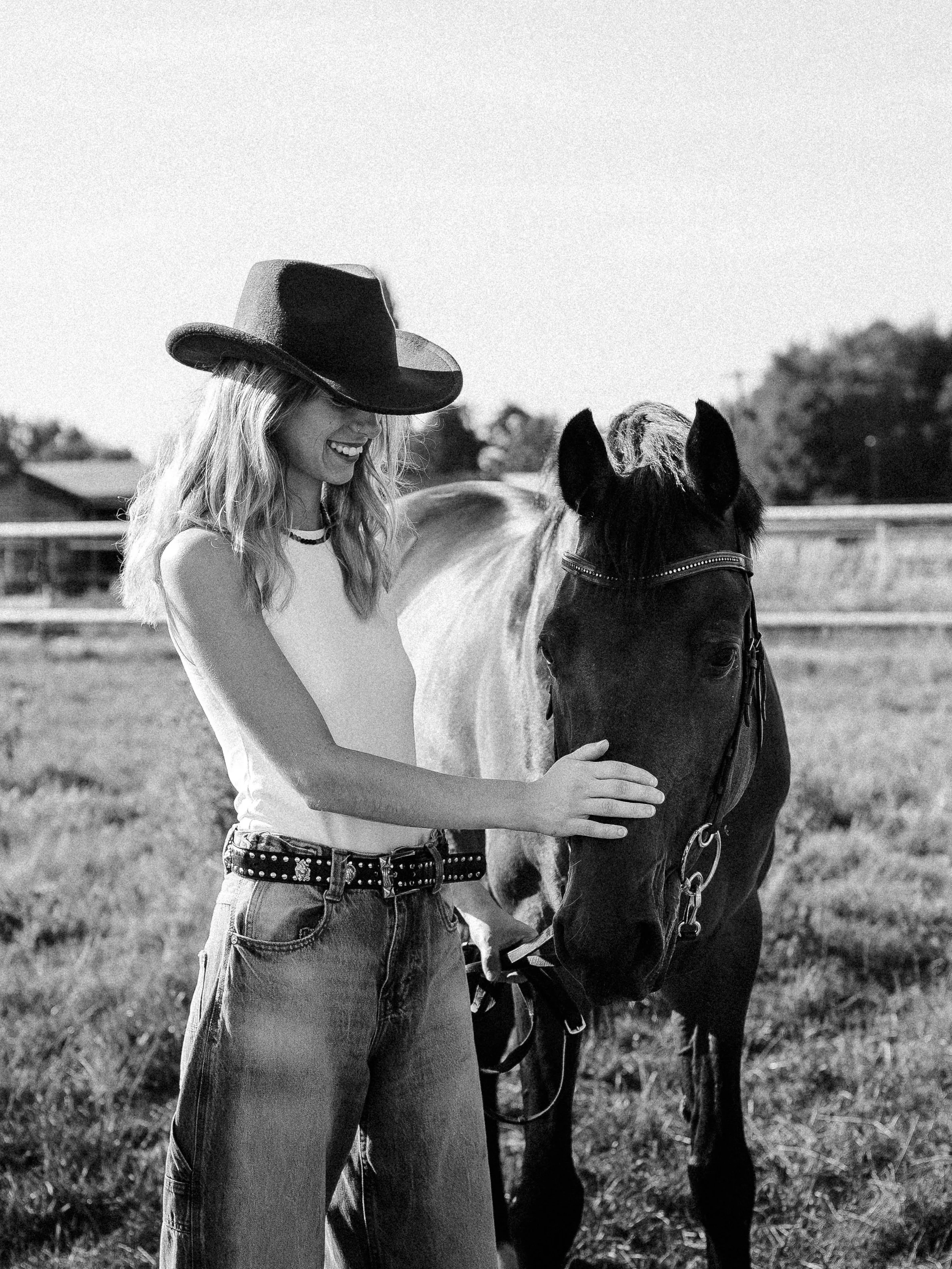 Une femme souriante avec un chapeau cowboy caresse un cheval dans un champ.