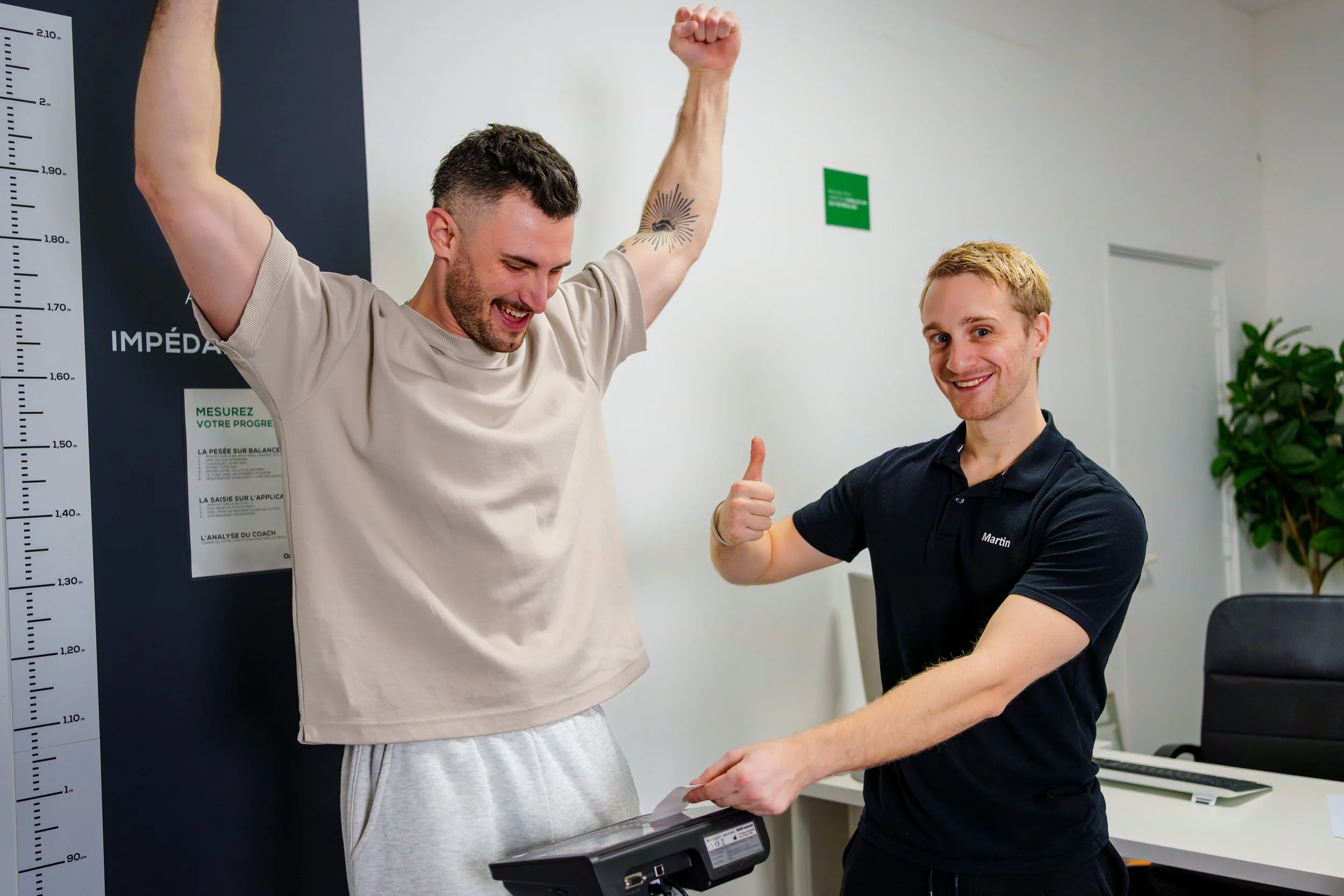 Deux hommes souriants, l'un avec les bras levés en signe de victoire, l'autre lui donnant un pouce en l'air, semblant célébrer une réussite dans un environnement de salle de sport ou de coaching.