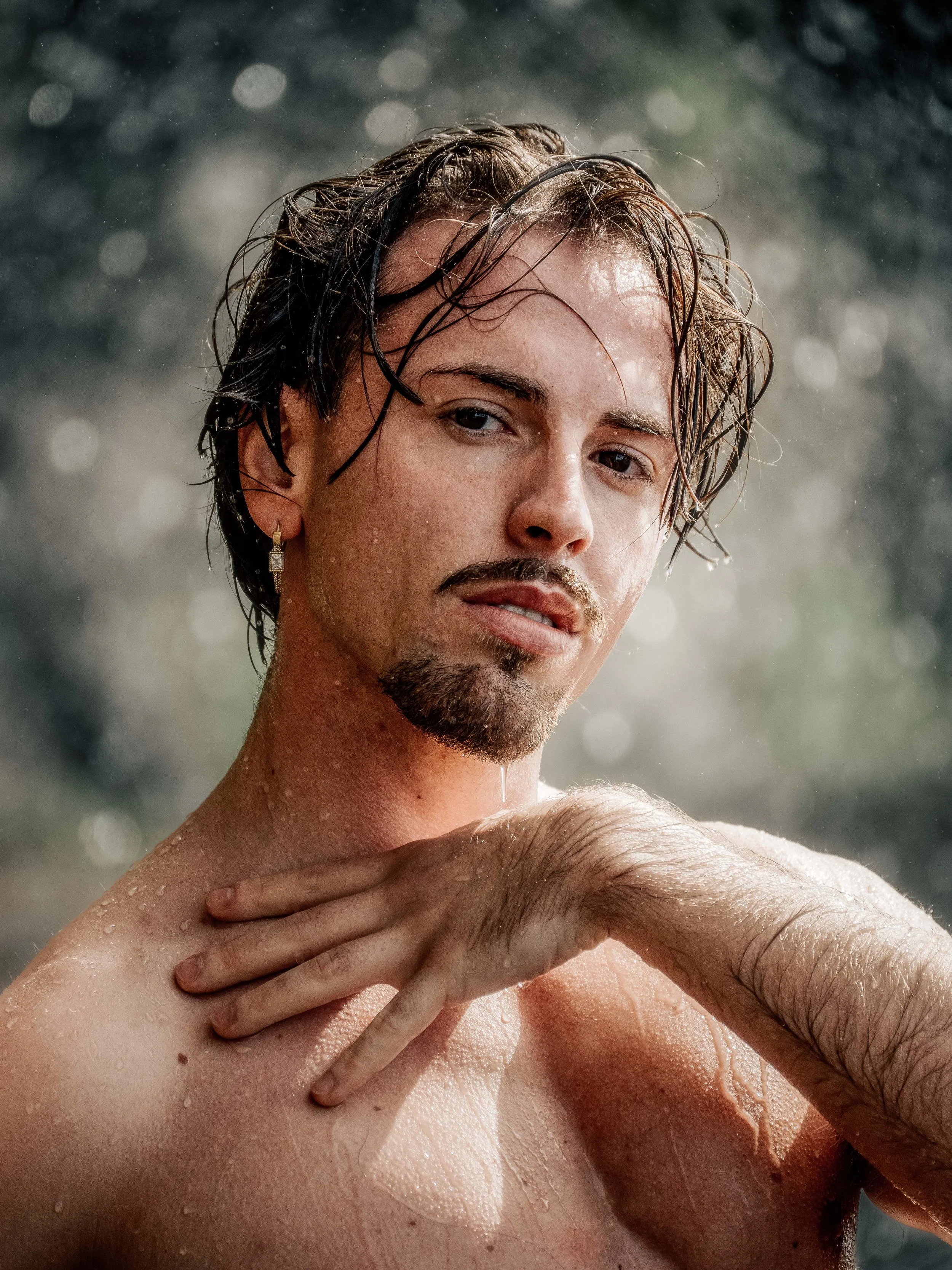 Jeune homme avec des cheveux mouillés, portant une boucle d'oreille, se tenant la tête, avec des gouttelettes d'eau et un fond flou.