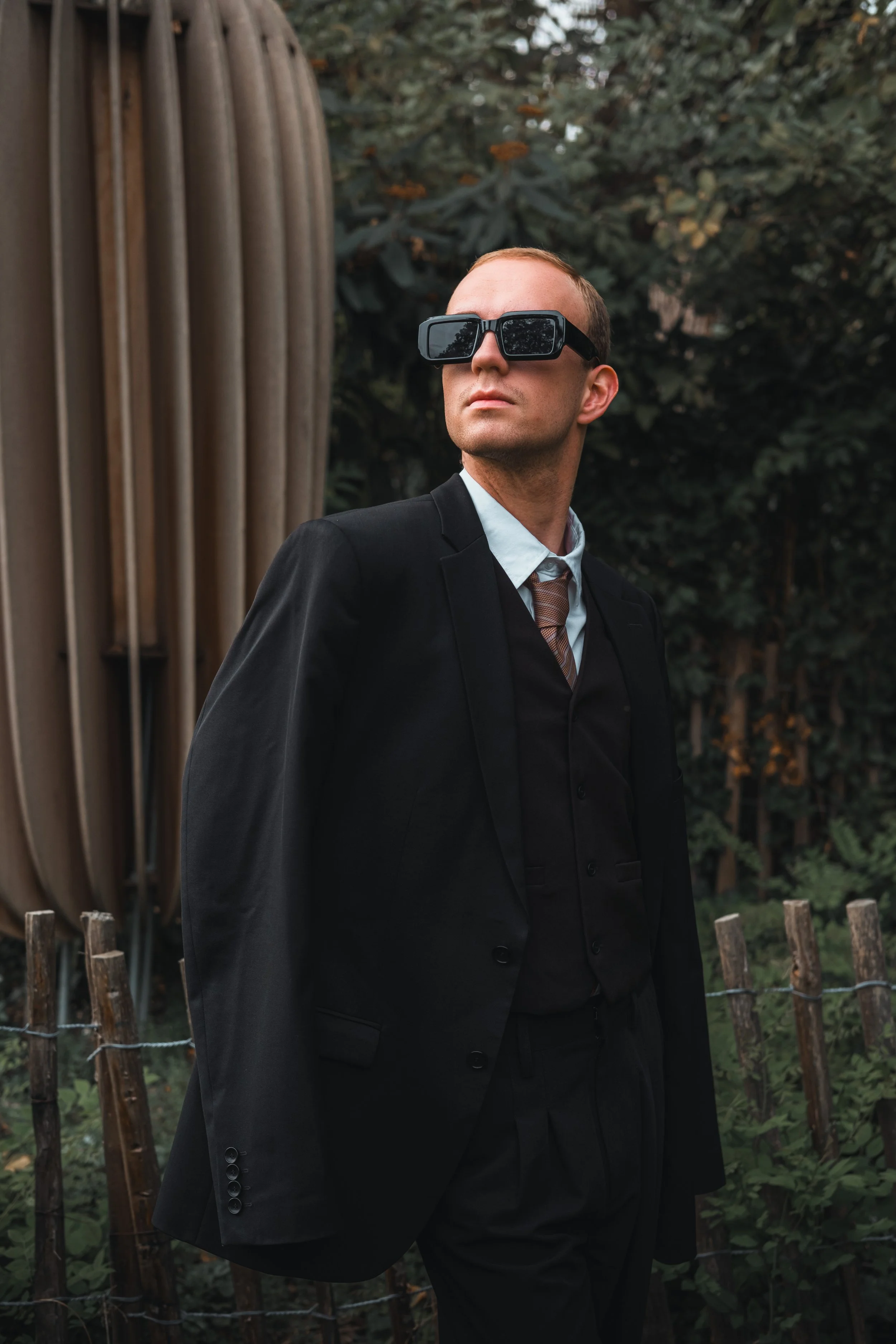 Homme en costume noir portant des lunettes de soleil, posé devant un décor naturel avec des arbres et une clôture en bois.