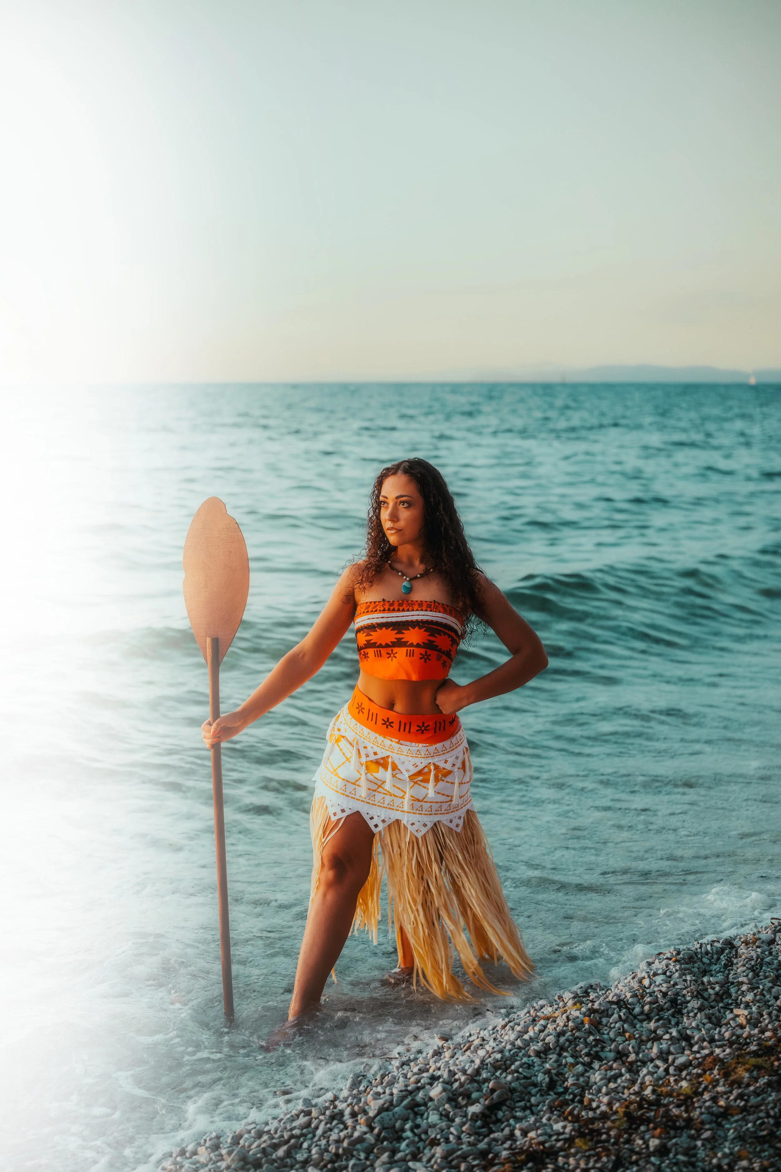 Femme en costume traditionnel hawaïen avec une pagaie en main, debout dans la mer sur une plage de galets, regardant vers la droite.