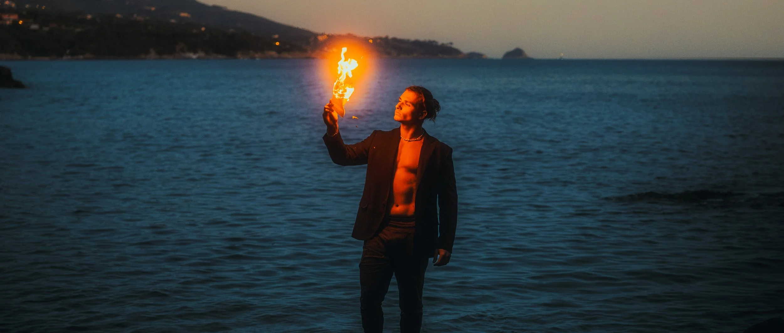 Homme debout près de la mer en soirée, tenant une torche enflammée.