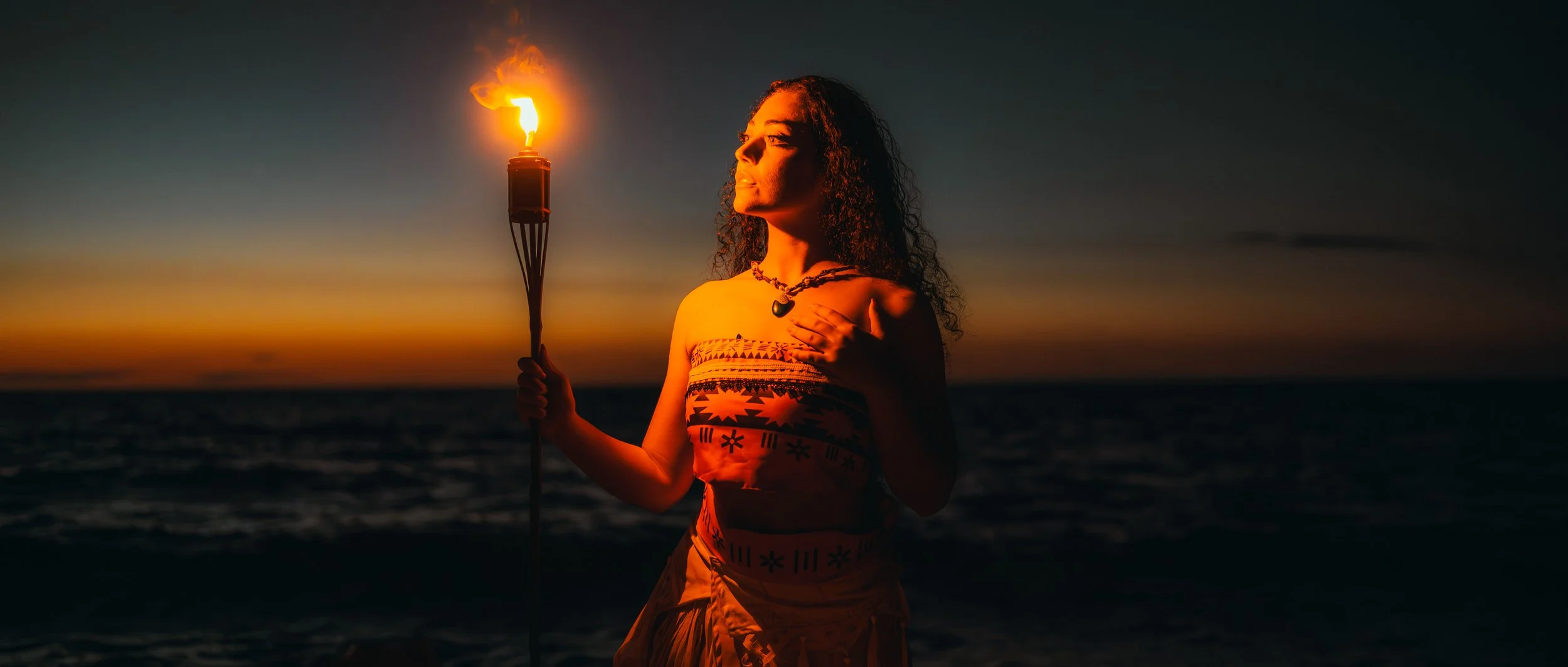 Une femme tient un bâton avec une torche en flamme sur une plage au coucher du soleil, portant un vêtement traditionnel et une colombe autour du cou.