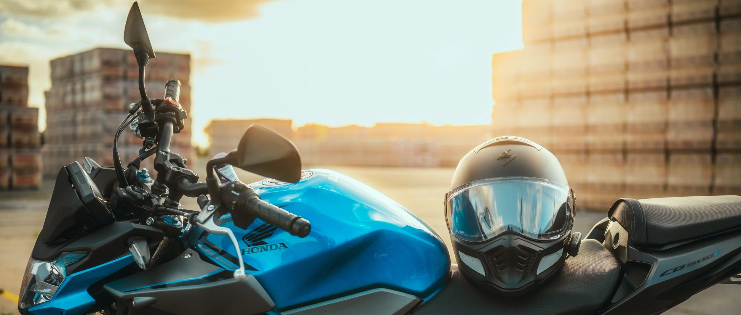 Une moto bleue avec un casque gris posé sur la selle dans un environnement en extérieur, avec un coucher de soleil en arrière-plan et des stacks de briques dans le fond.