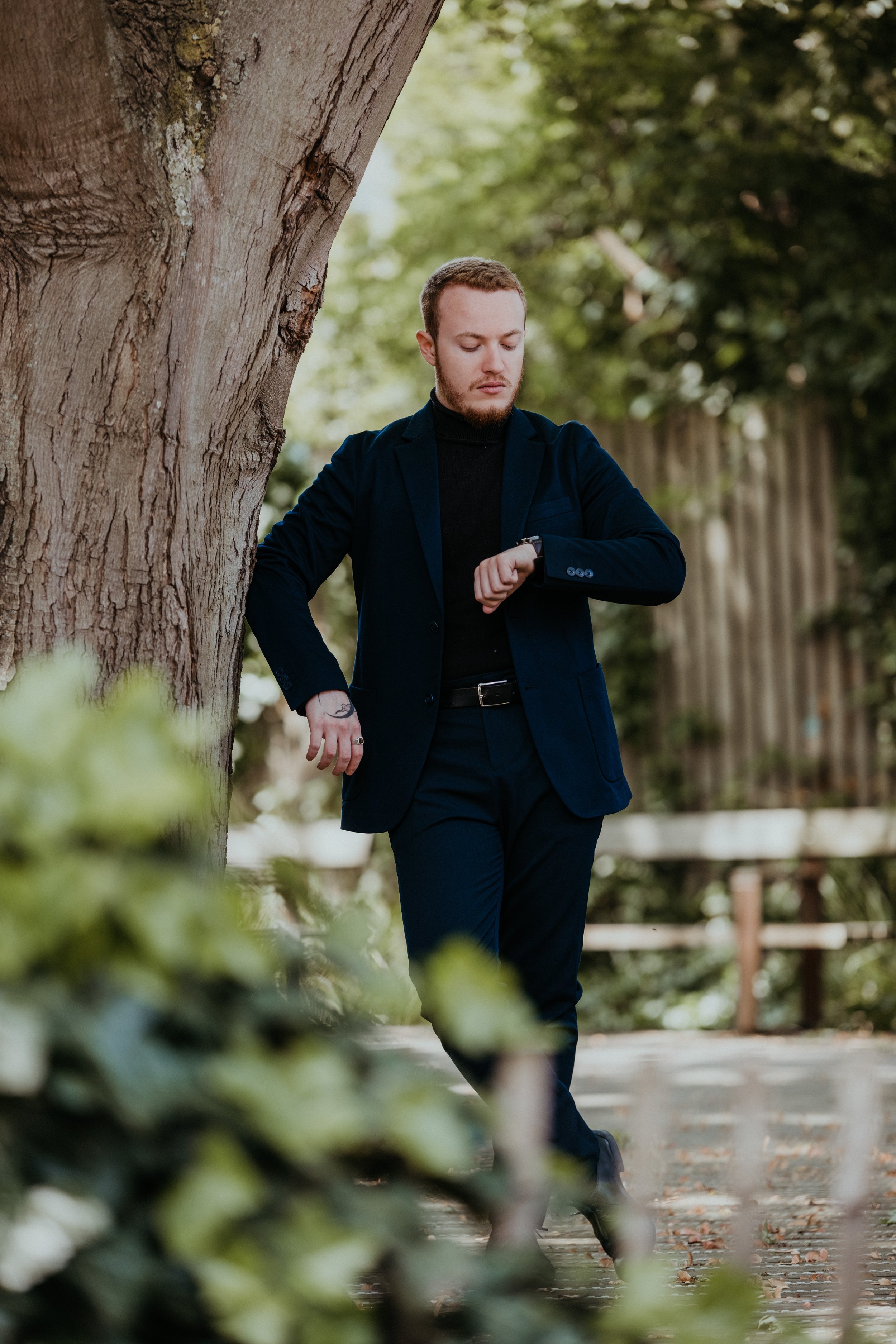 Jeune homme en costume noir à l'extérieur, regardant sa montre, près d'un arbre, avec un fond de nature verdoyante et un chemin en pierre