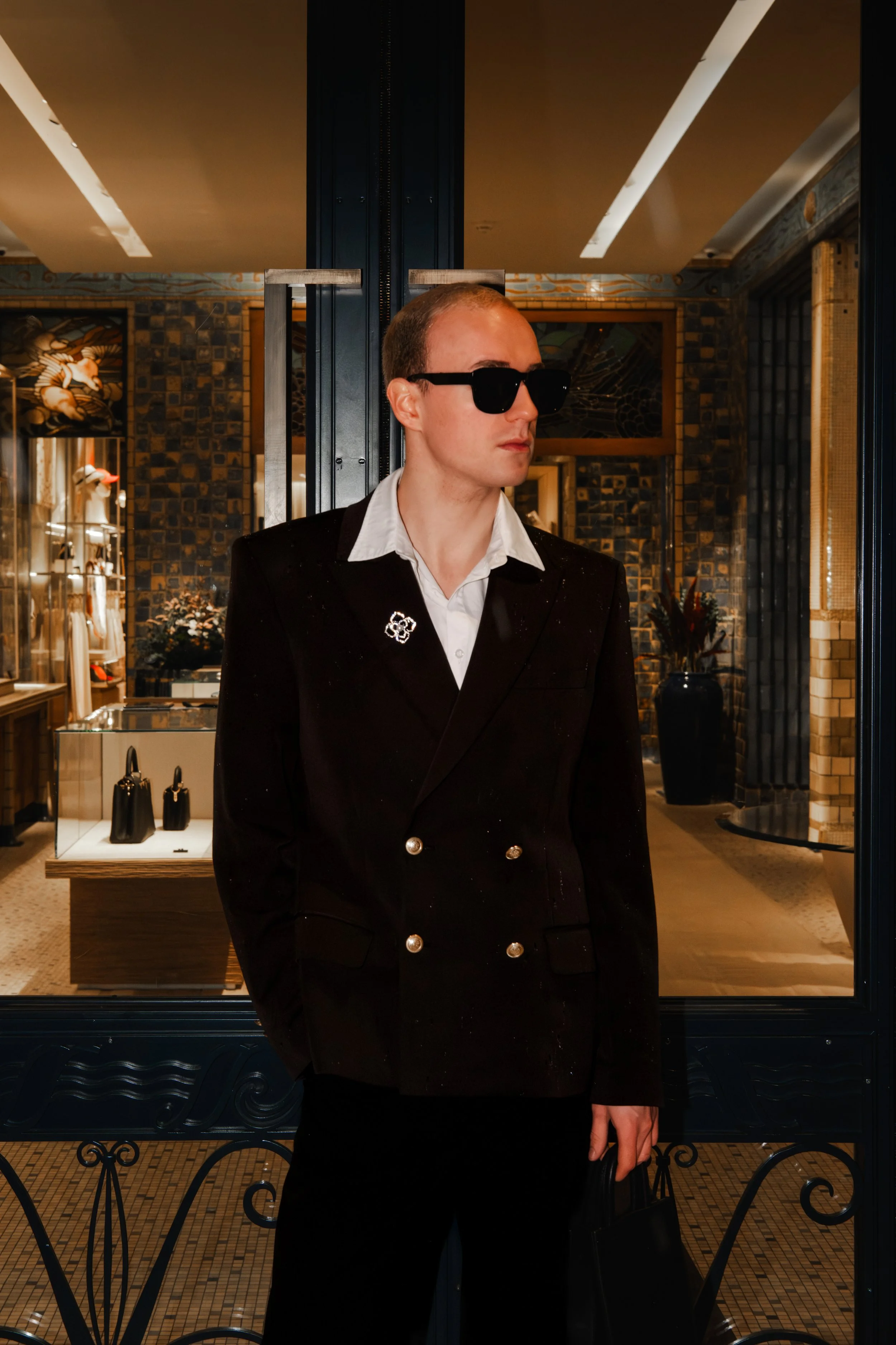 Homme portant un costume noir avec des boutons dorés, portant des lunettes de soleil noires, se tenant devant une vitrine dans une boutique de mode.