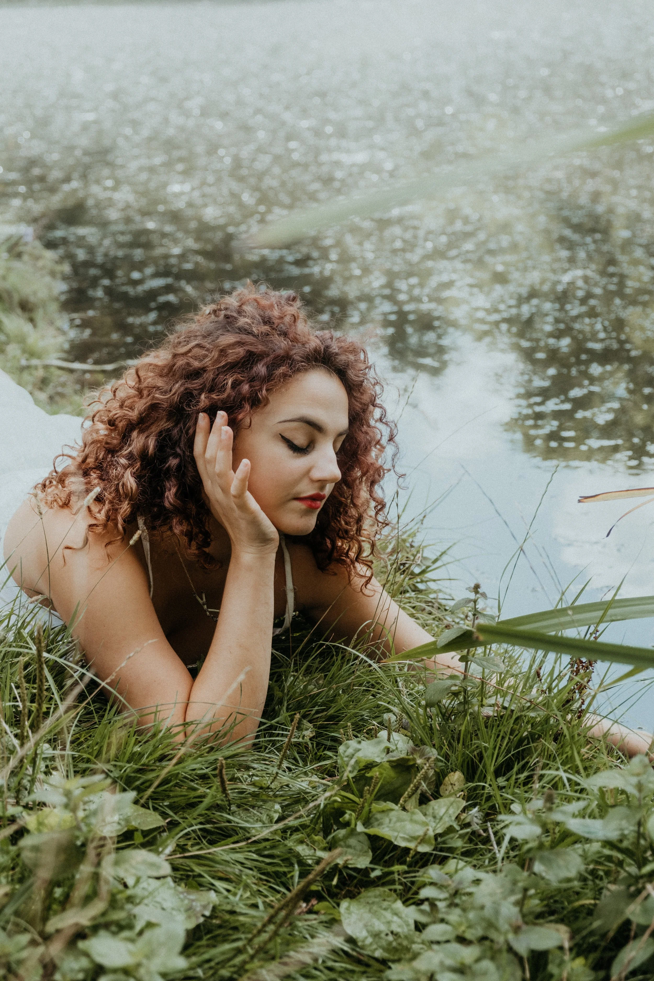Jeune femme aux cheveux bouclés allongée sur l'herbe à côté d'un corps d'eau, les yeux fermés, en train de se détendre dans la nature.